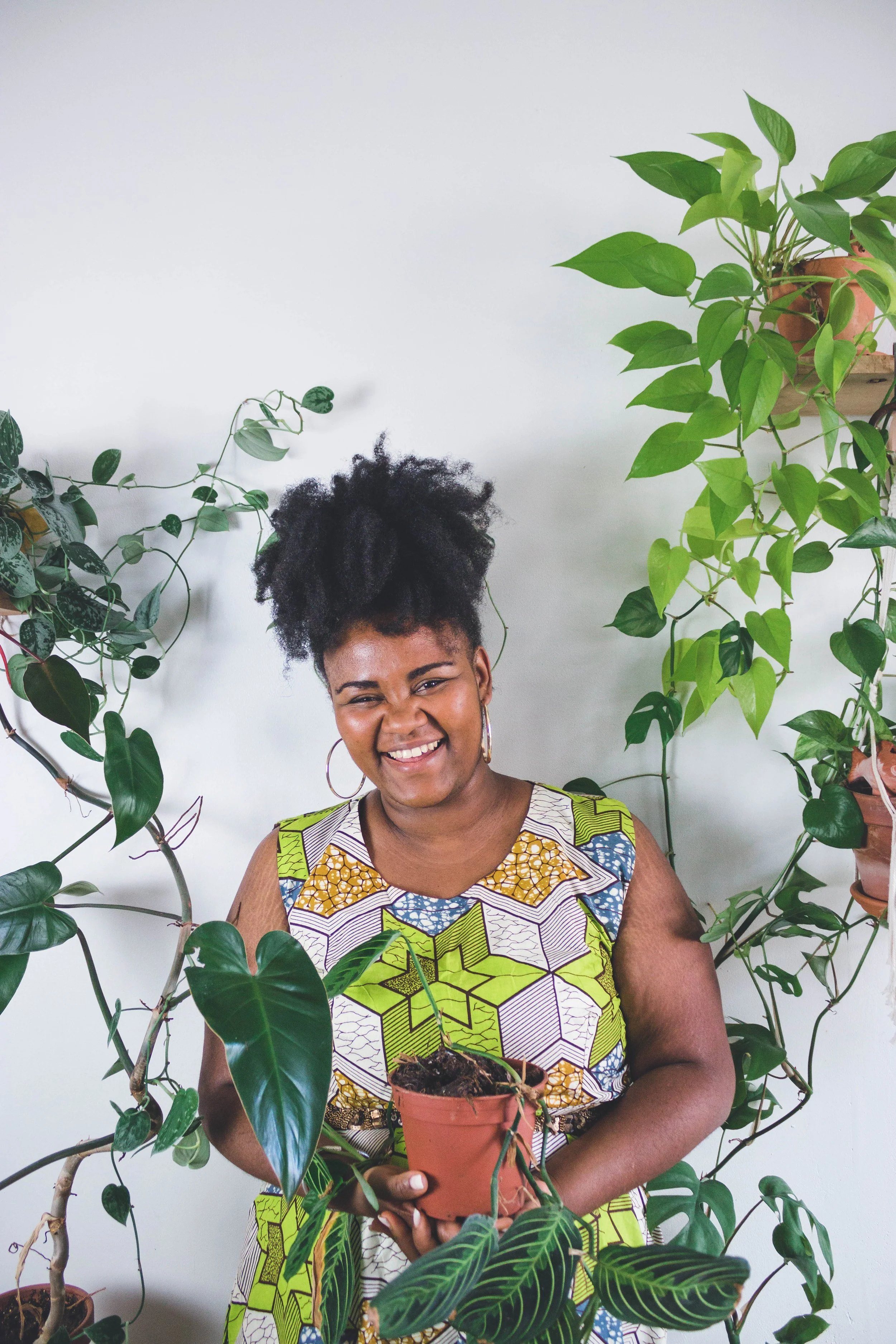 Femme souriante tenant un pot de plante d'intérieur avec plusieurs plantes vertes autour, debout devant un mur blanc.