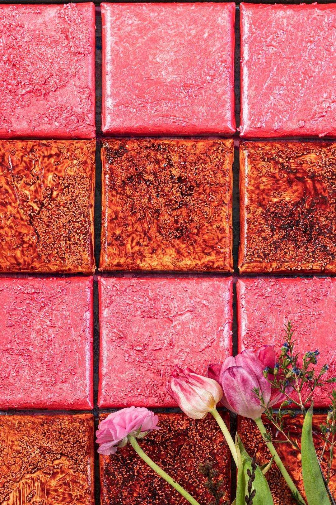 A top-down view of nine pink and brown square cakes arranged in a 3x3 grid, with pink flowers and greenery in the bottom right corner.