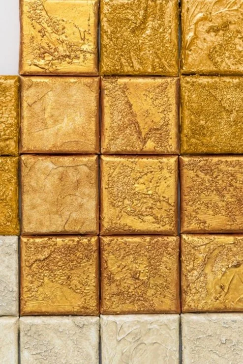 Close-up of a tiled wall with square tiles in shades of gold and beige, featuring textured and marbled patterns.