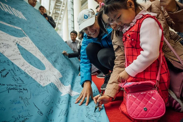A young girl with a pink bag signs a large blue banner covered in signatures, assisted by an adult wearing a blue jacket and a white cap. Several other people are in the background inside a building.
