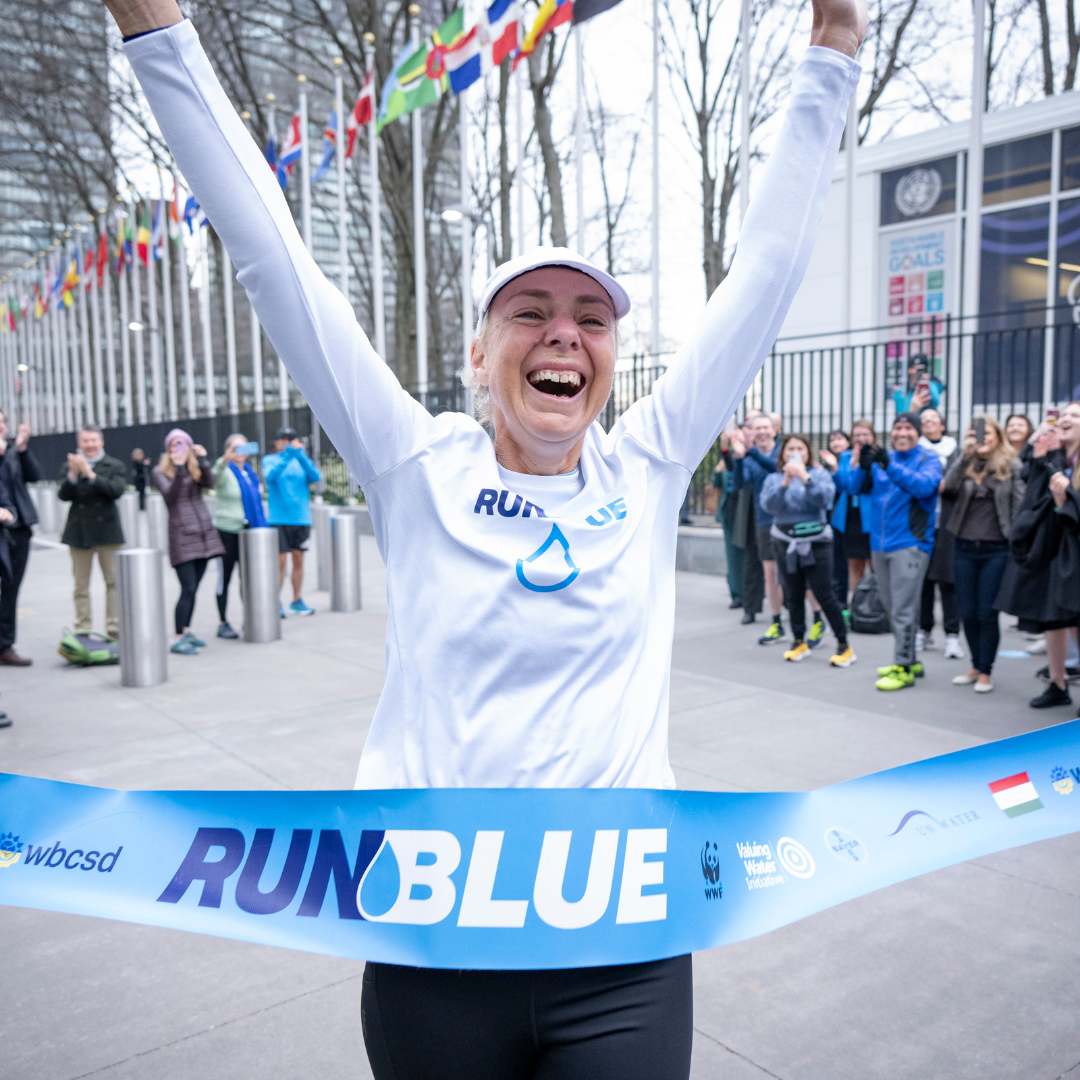 Runner celebrating crossing the finish line at an outdoor event, surrounded by cheering crowd, holding a blue "RUN BLUE" banner.