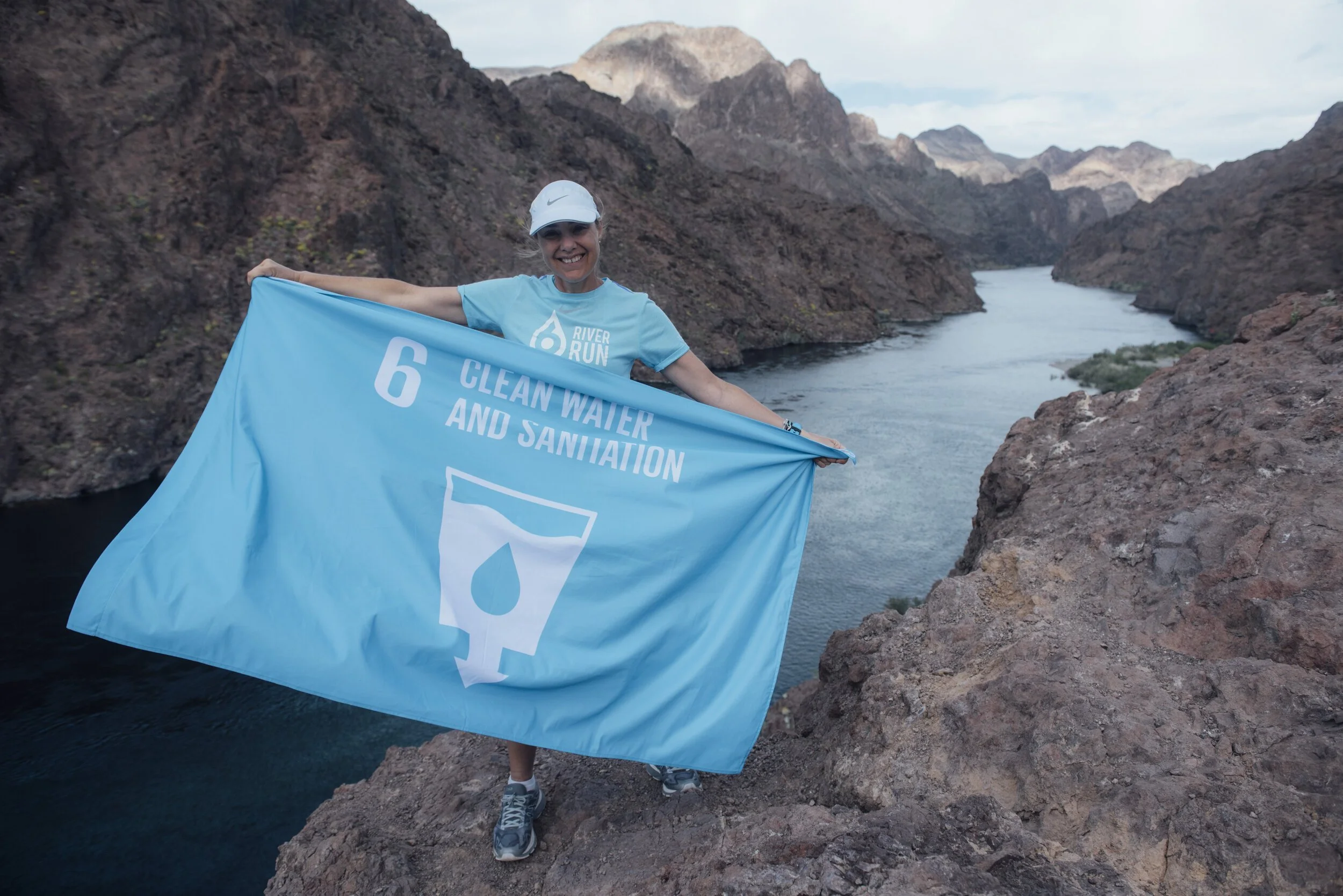 Person holding a blue flag with the text '6 Clean Water and Sanitation' near a river in a rocky mountainous area.