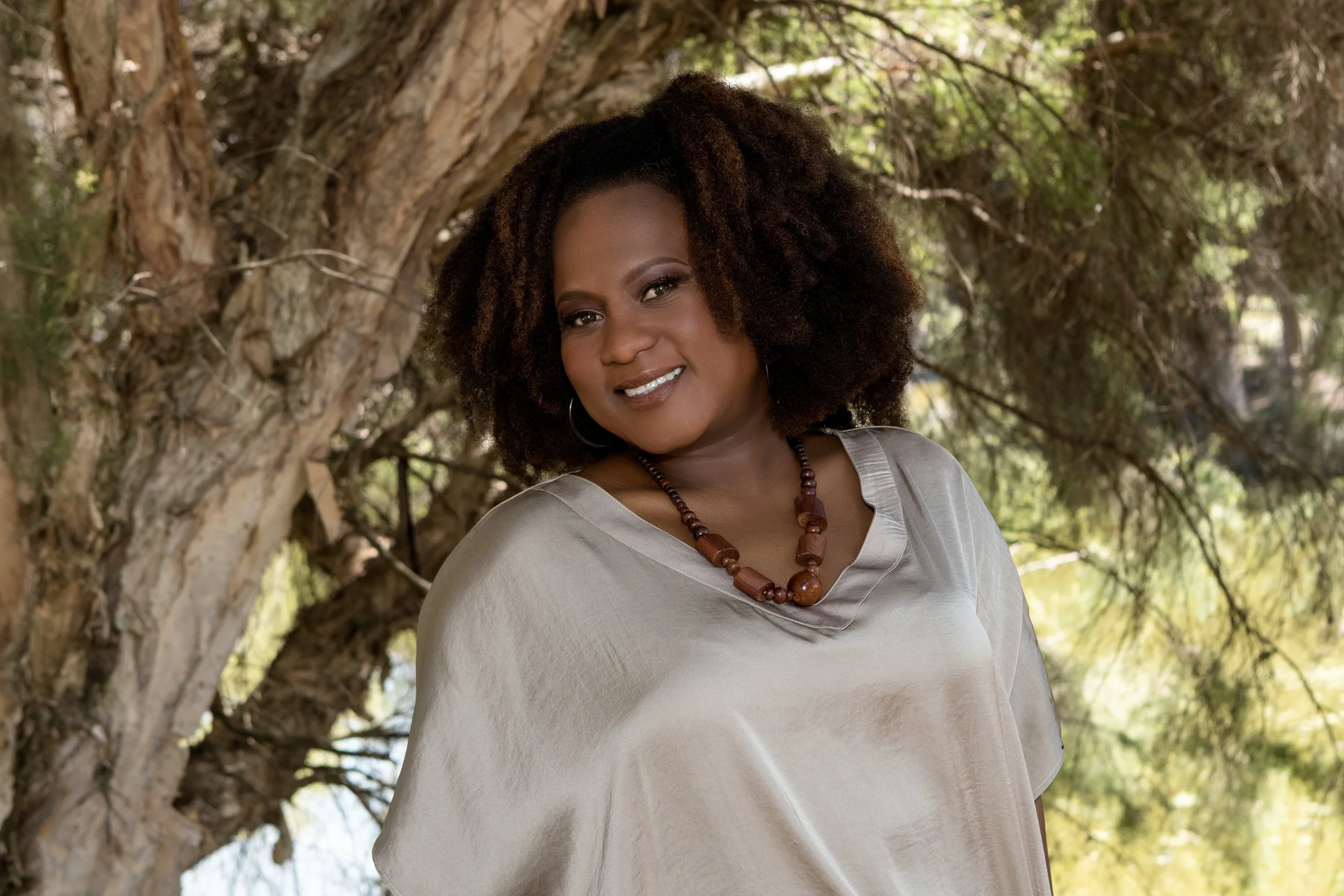 A woman with dark skin and curly hair, wearing a beige satin blouse and a large wooden beaded necklace, standing outdoors in front of a tree with green foliage.