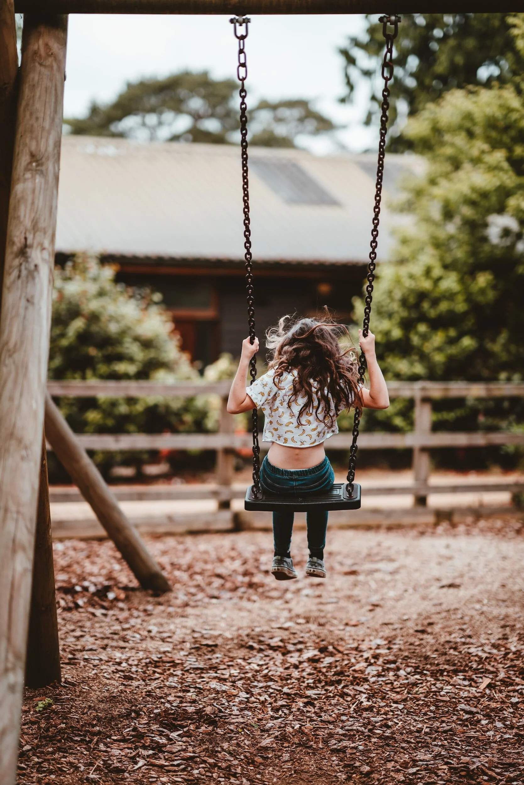 A girl with curly hair swinging alone on a swing set outdoors with trees and a building in the background.