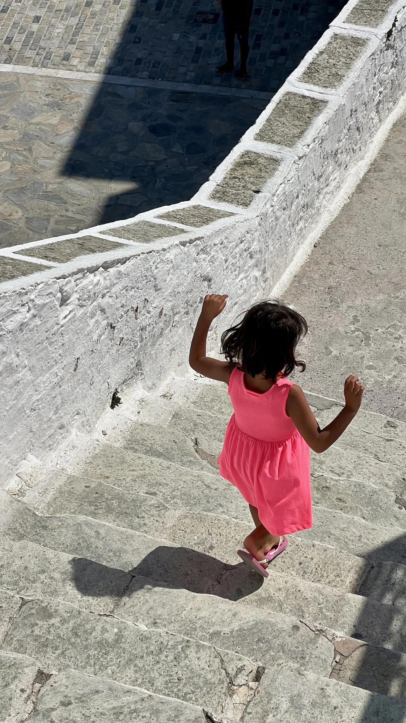 A young girl in a pink dress and flip-flops runs down stone steps outside, with a white stone wall on one side and shadows cast over the area.