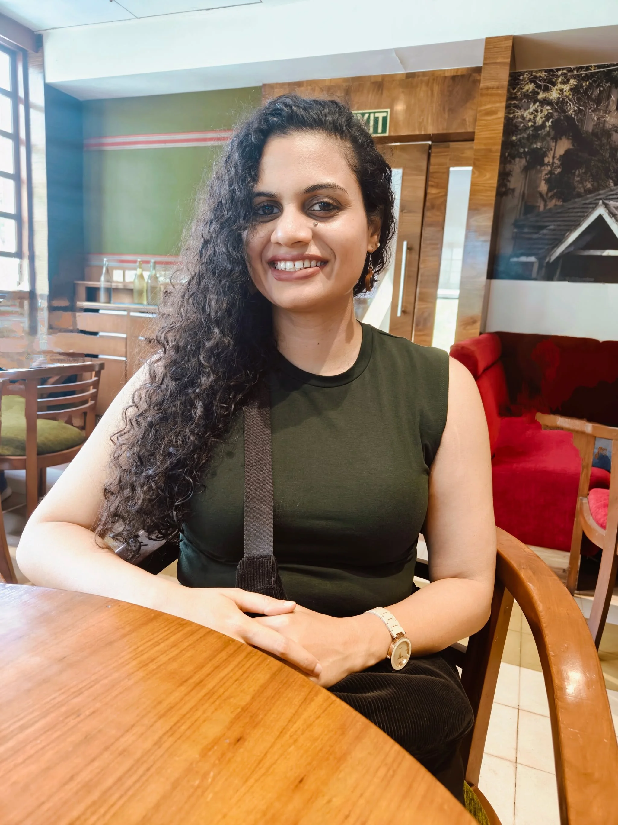 A woman with curly dark hair, wearing a sleeveless dark green top, seated at a wooden table in a cafe, smiling at the camera.