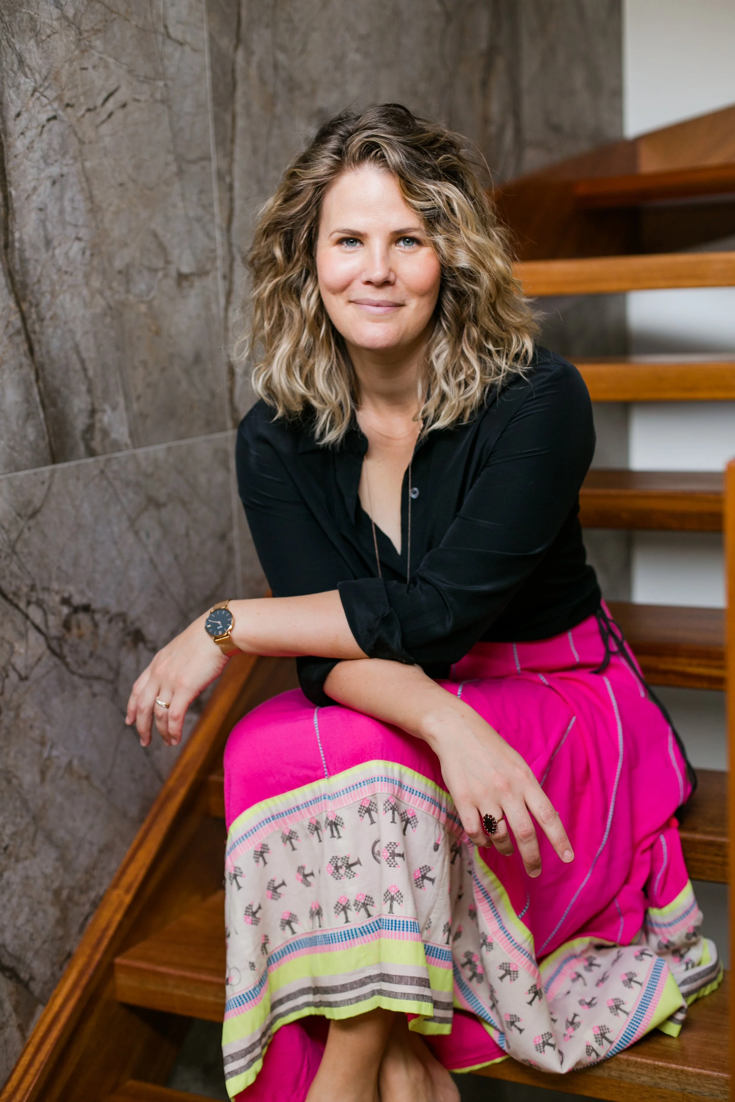 A woman with curly blonde hair sits on wooden stairs against a stone wall, wearing a black blouse and a colorful patterned skirt, smiling at the camera.