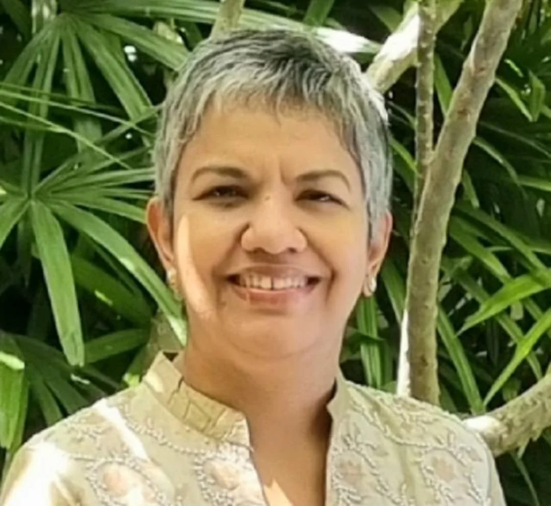 A smiling woman with short gray hair, wearing a light-colored traditional Asian-style shirt, standing outdoors with green leafy plants in the background.