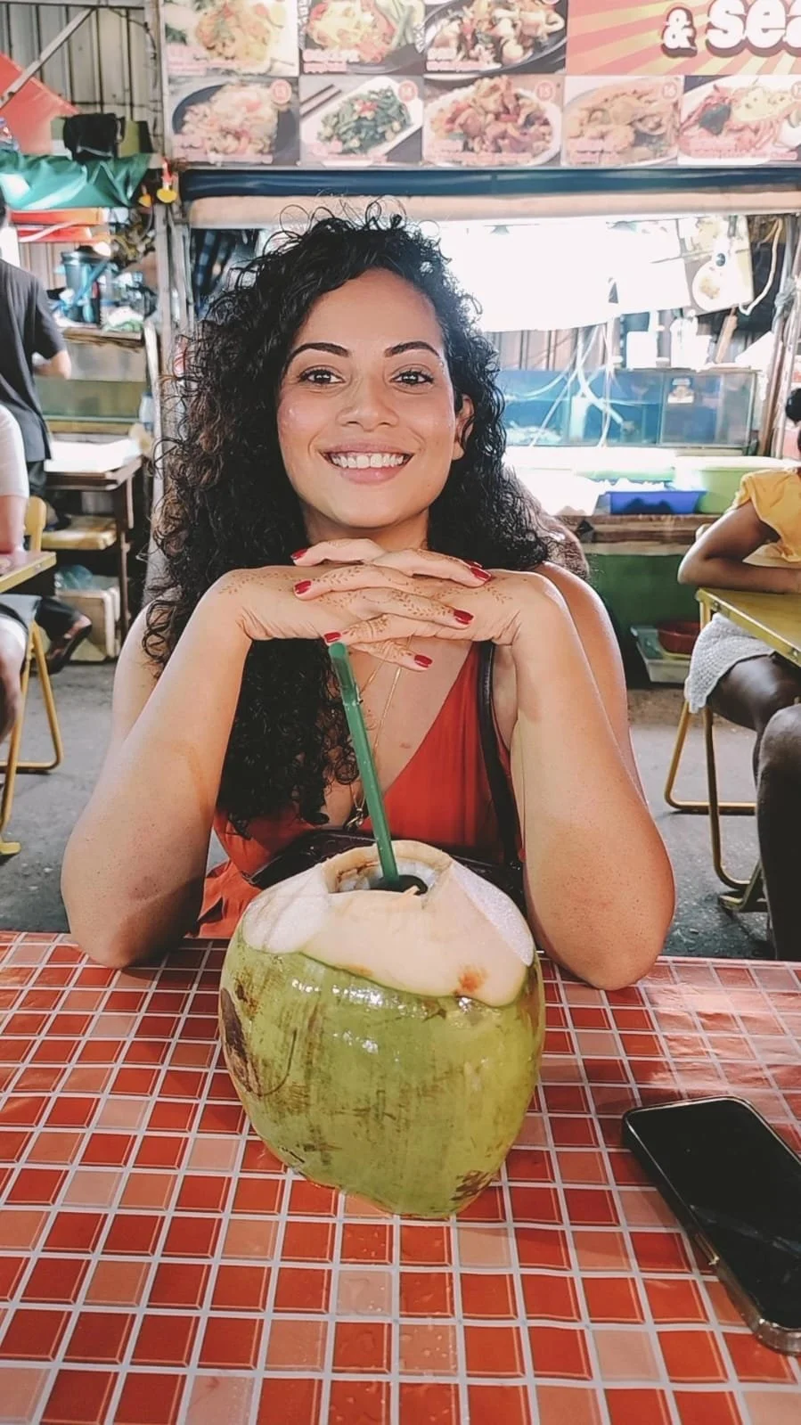 A woman with curly black hair, wearing a sleeveless orange top, sitting at a table in a restaurant, smiling, with a large green coconut drink with a straw in front of her. The table has a red checkered surface.