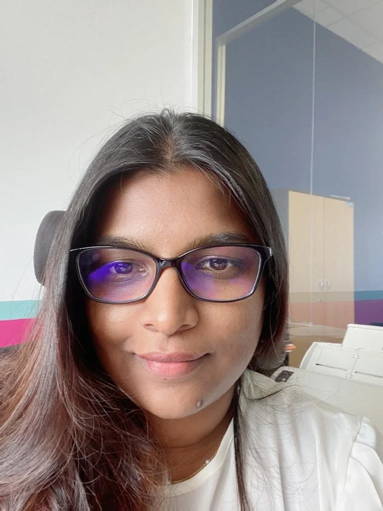 Close-up of a woman wearing glasses with purple-tinted lenses, smiling indoors with office furniture and a blue wall in the background.