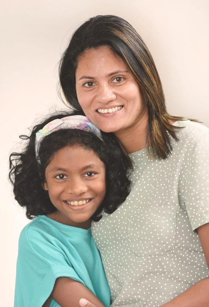 A woman and a young girl smiling and hugging in front of a plain background.