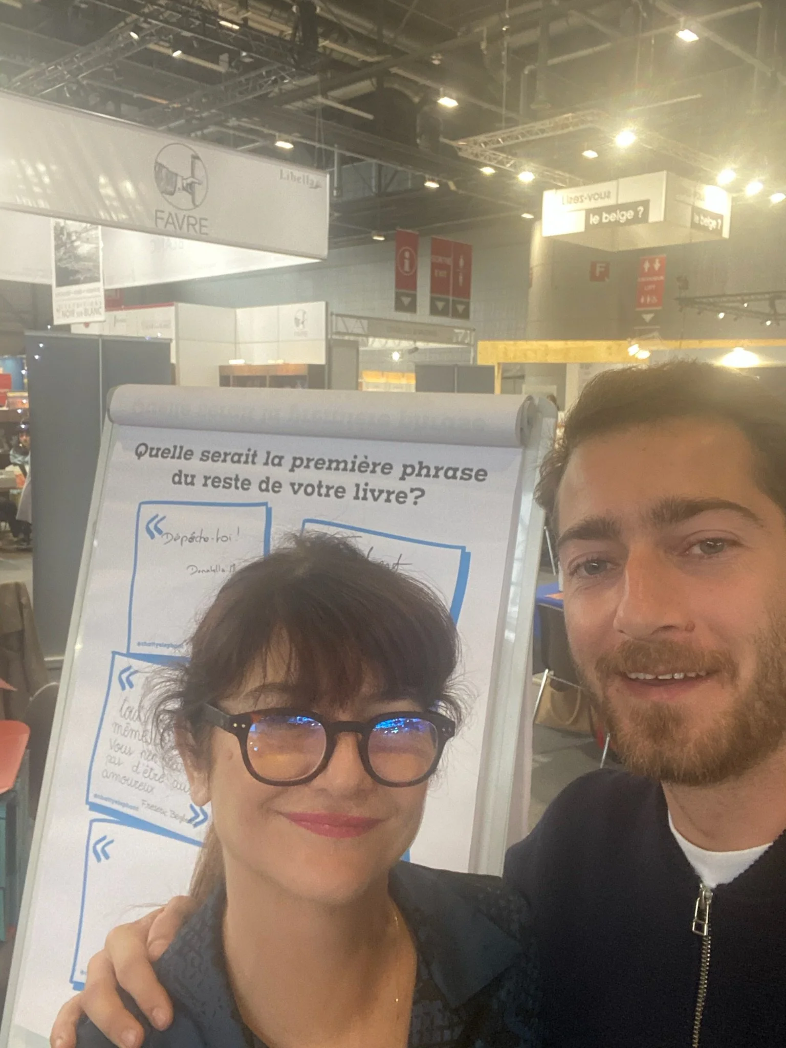 Deux personnes prennent un selfie devant un tableau blanc avec des questions en français, dans un centre commercial ou une foire. La femme porte des lunettes noires et des vêtements noirs, et l'homme a une barbe et porte une veste noire.