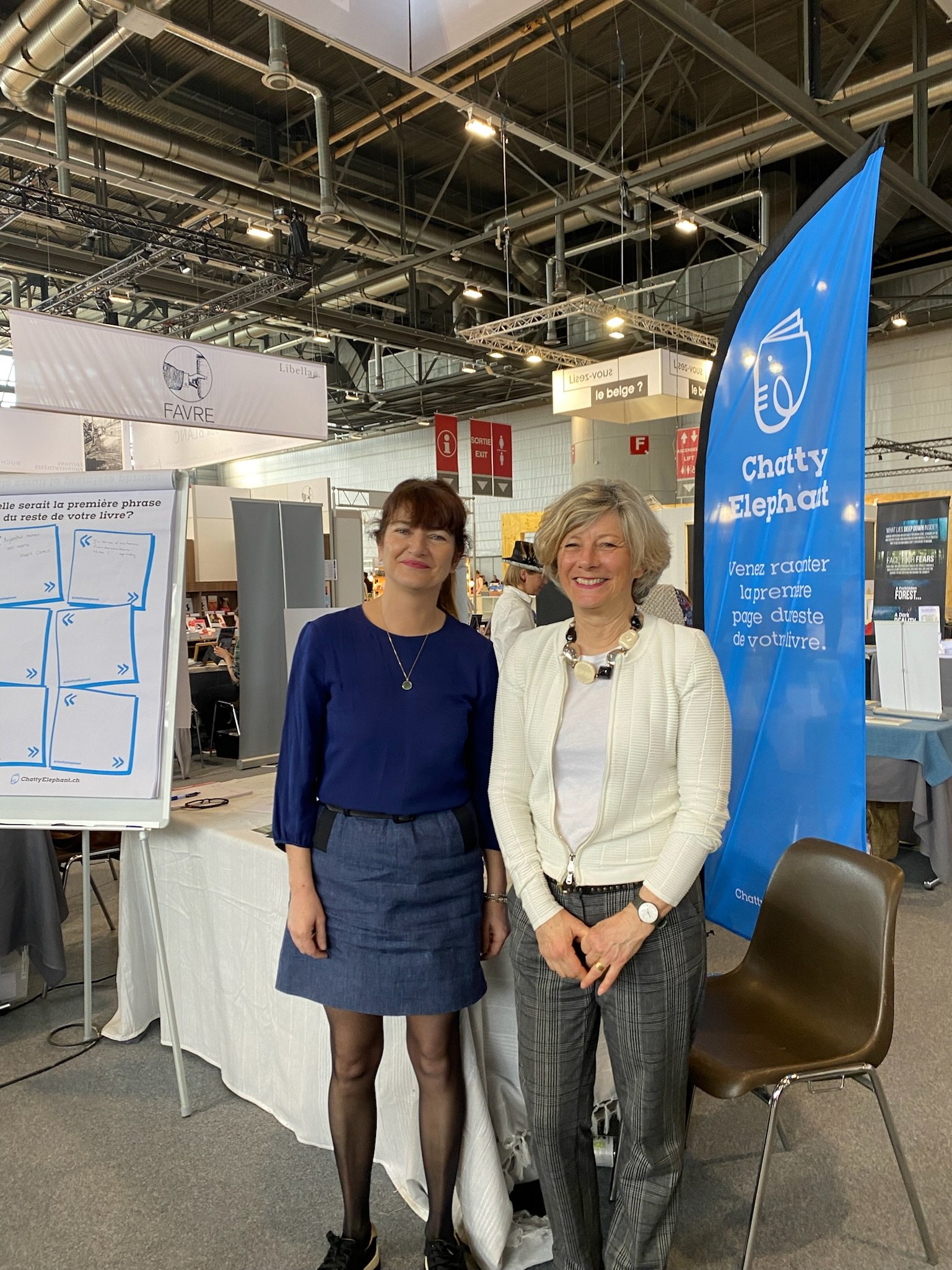 Deux femmes debout côte à côte dans un salon ou une foire, à côté d'une bannière bleue avec un éléphant blanc et le texte "Chatty Elephat". Elles sourient, l'une porte une robe bleu foncé et l'autre une veste blanche avec un pantalon à carreaux.