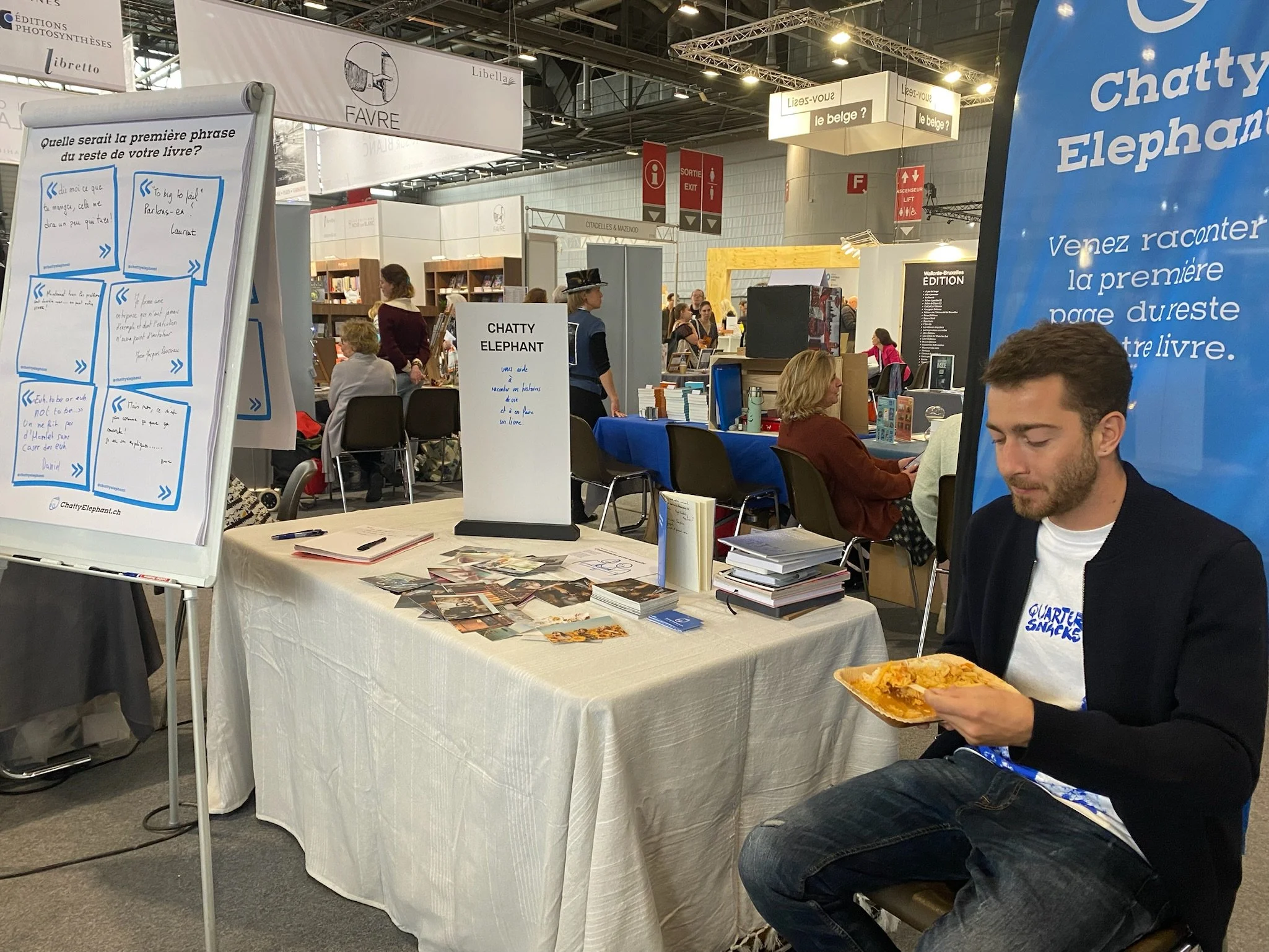Un homme assis à une table de dédicaces lors d'une foire du livre, mangeant un repas, avec d'autres personnes lisant ou discutant dans le fond.
