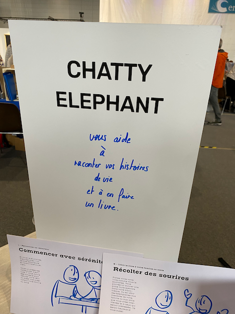 Panneau blanc avec le texte "Chatty Elephant" en haut, et une écriture manuscrite bleue disant "vous aide à rencontrer vos histoires de vie et à en faire un livre", avec deux feuilles de papier dessous contenant de petits dessins et textes.