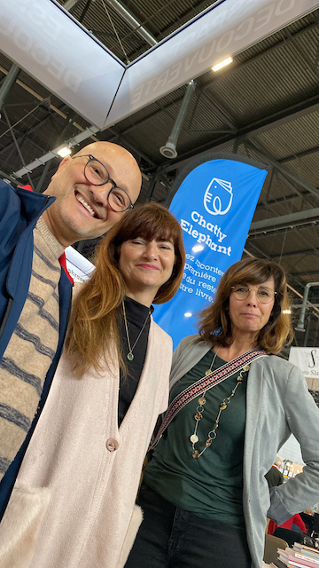Trois personnes posent pour une photo dans un centre d'exposition, avec un drapeau bleu de l'association Charité Elephant en arrière-plan.