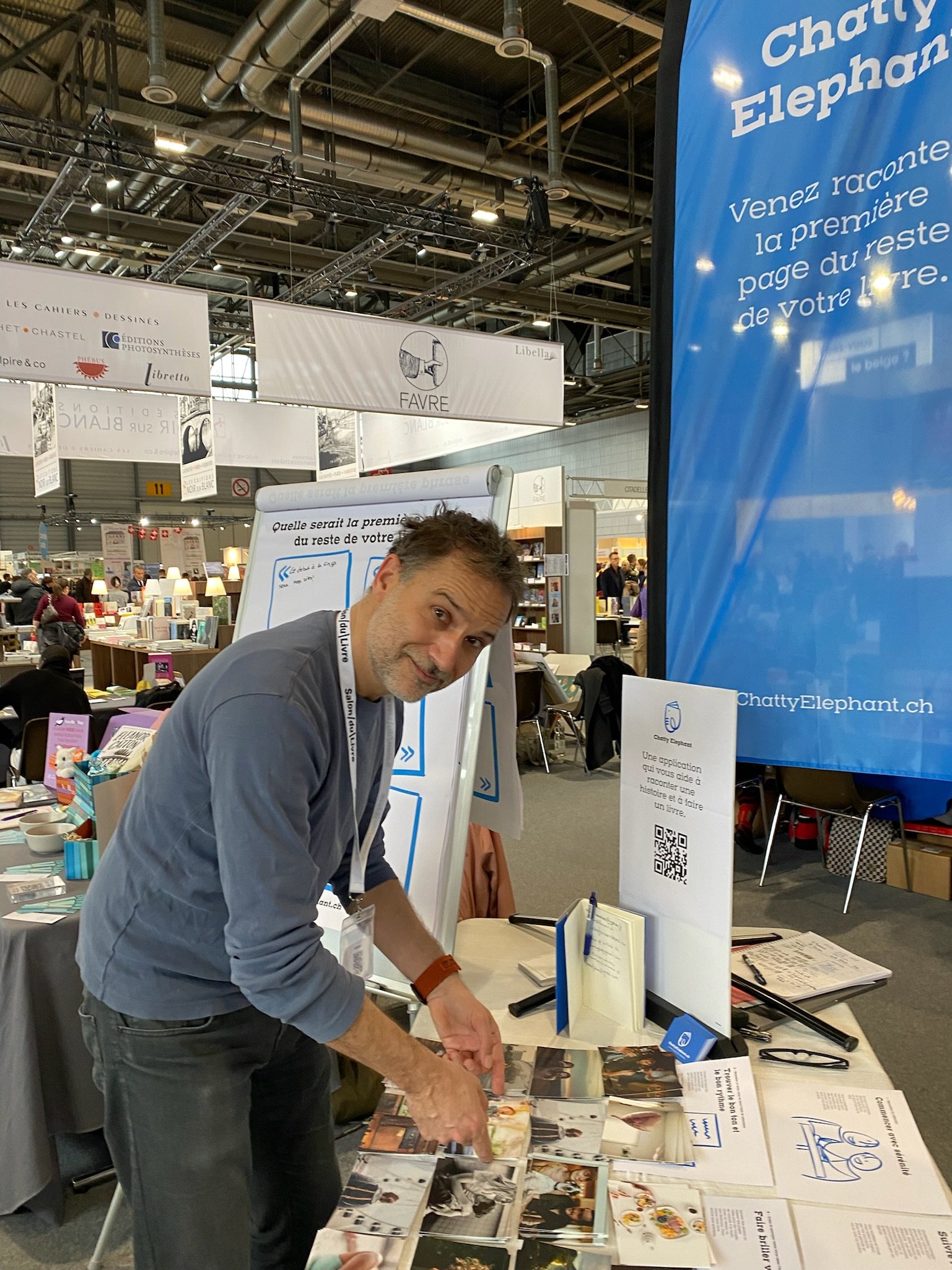 Un homme debout à un stand dans une foire ou salon, feuilletant des brochures ou des photos, avec des banners et des stands remplis de livres et de matériel en arrière-plan.