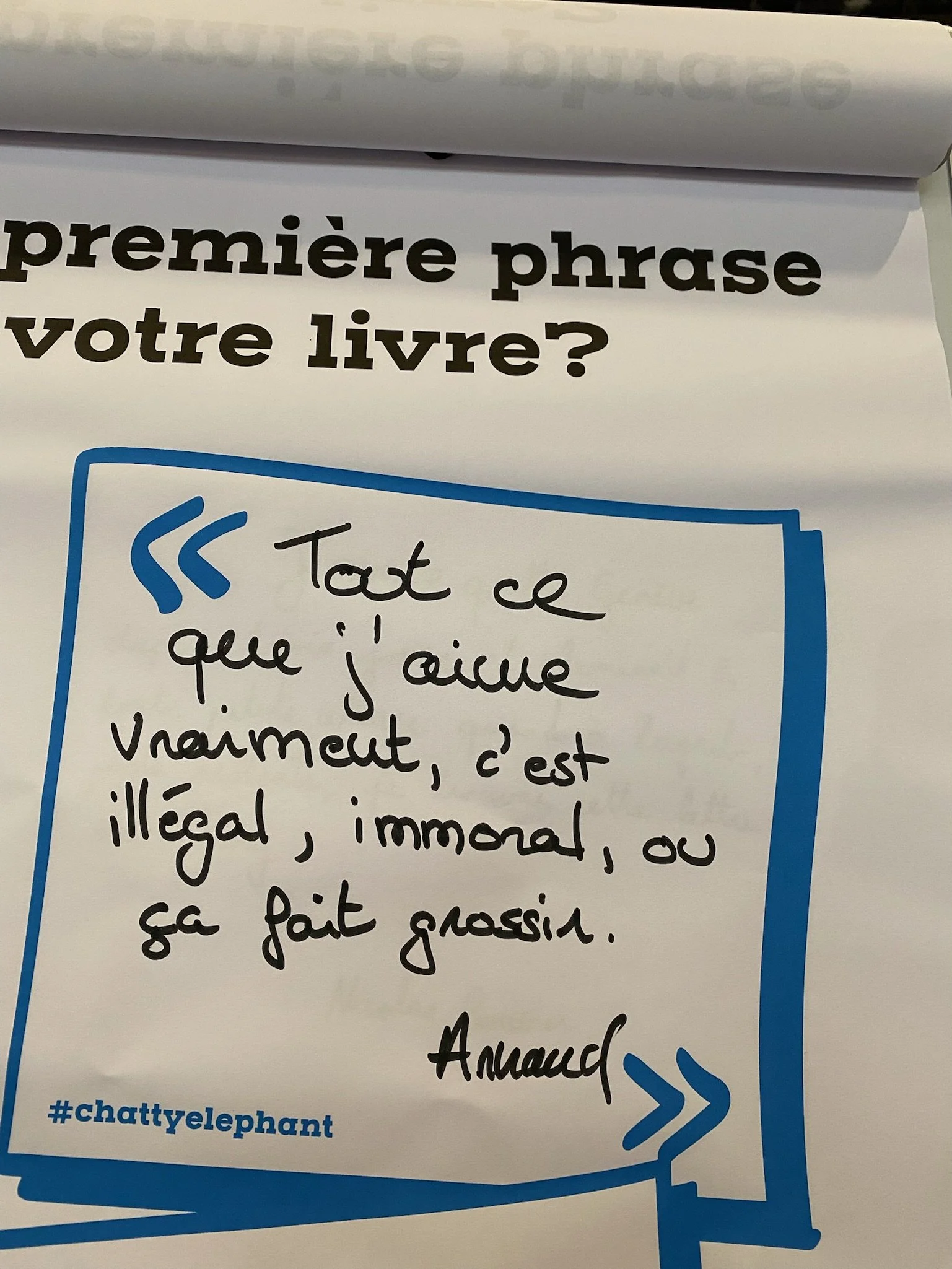 Une feuille de papier avec une question en français : "Quelle est la première phrase de votre livre ?" et une réponse écrite manuscrite : "Tout ce que j'aime vraiment, c'est illégal, immoral, ou ça fait grossir." La feuille comporte aussi le mot-clé 