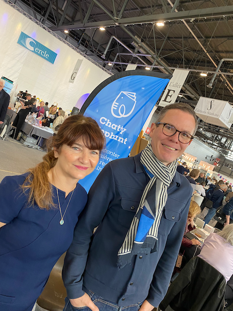 Deux personnes posent pour la photo à un événement, un homme et une femme, devant un stand avec un drapeau bleu et un logo d'un livre, dans une grande salle d'exposition.