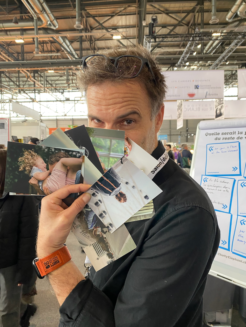 Un homme tient des photos devant son visage dans ce que semble être un salon ou une exposition. Il porte des lunettes sur la front et a un bracelet orange à son poignet gauche.