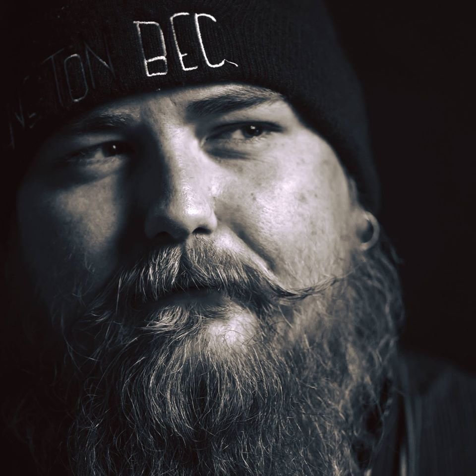 Un homme avec une barbe et une moustache, portant un bonnet noir avec l'inscription 'ON DEC' dans une lumière tamisée.