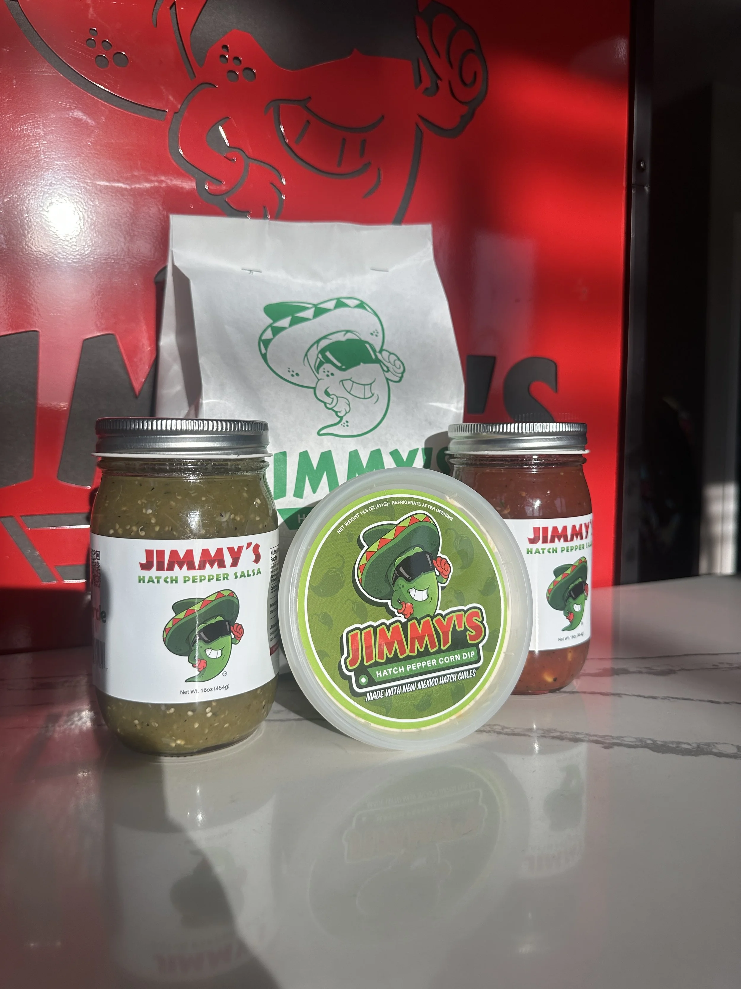 Three jars of Jimmy's Hatch Pepper salsa and corn dip on a white surface with a red Jimmy's shopping bag in the background.
