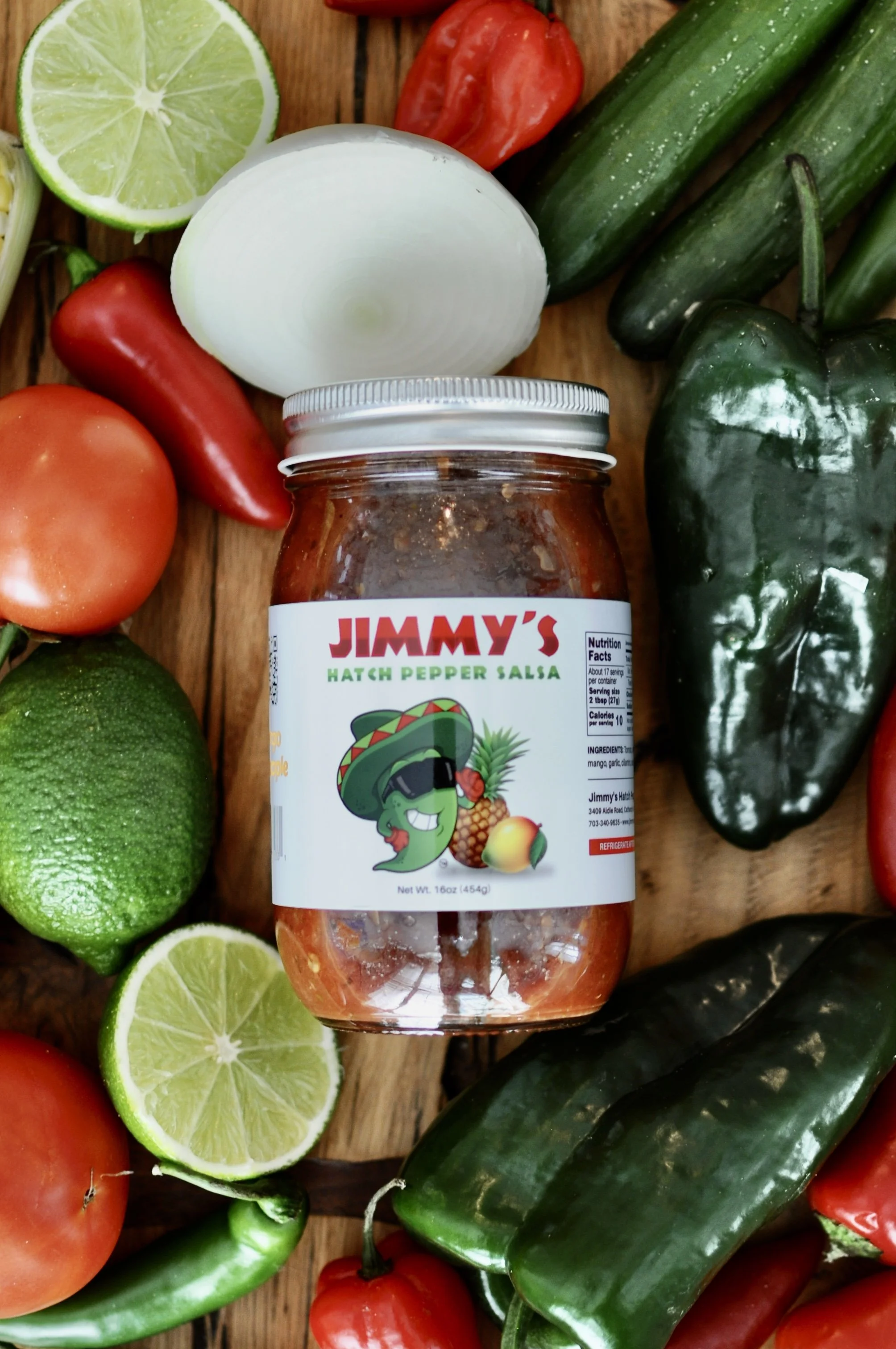 A jar of Jimmy's Hatch Pepper Salsa surrounded by various fresh vegetables and fruits, including tomatoes, cucumbers, onions, limes, and peppers, on a wooden surface.