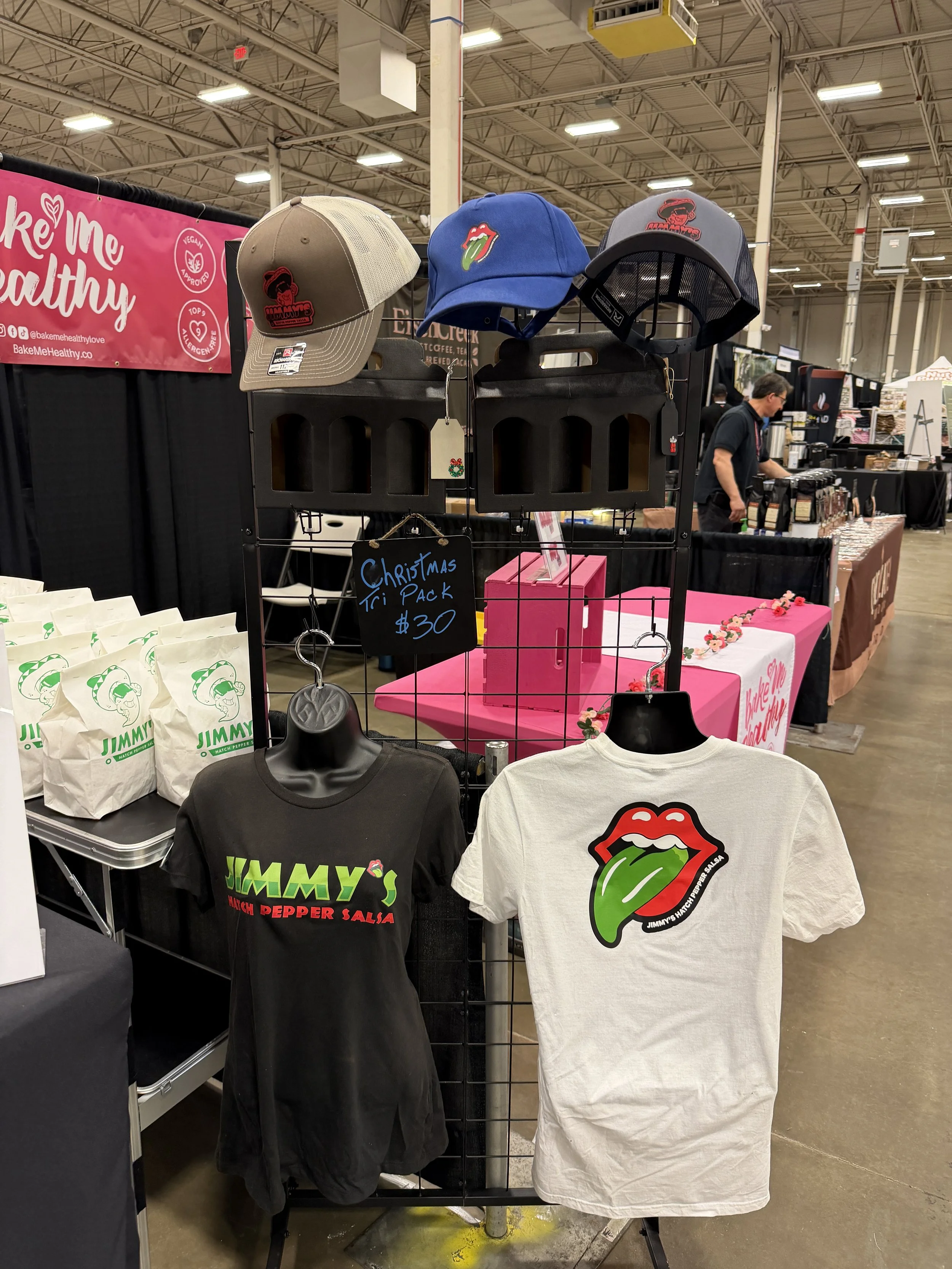 Display of Jimmy's hot pepper salsa merchandise at a market booth, featuring hats, T-shirts with a tongue and lips logo, and a sign advertising a Christmas gift bundle 
