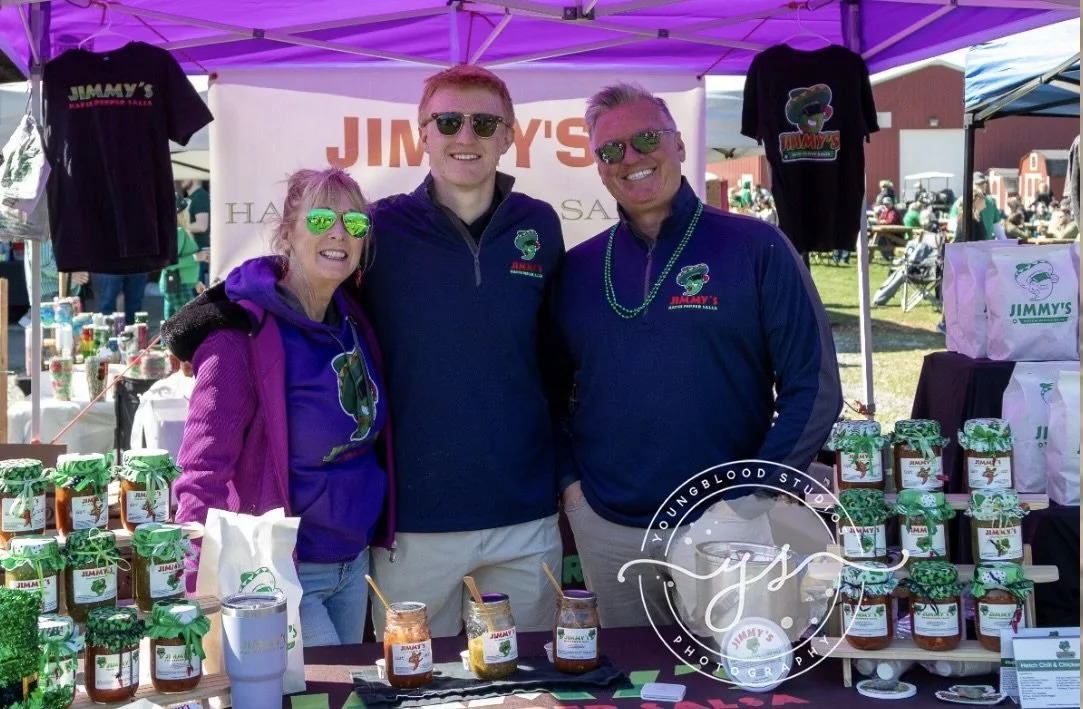 Three people standing under a tent at an outdoor market booth, smiling at the camera. The booth displays jars of jam with green and white labels, t-shirts with the Jimmy's logo, and a banner in the background. The people wear sunglasses and casual cl
