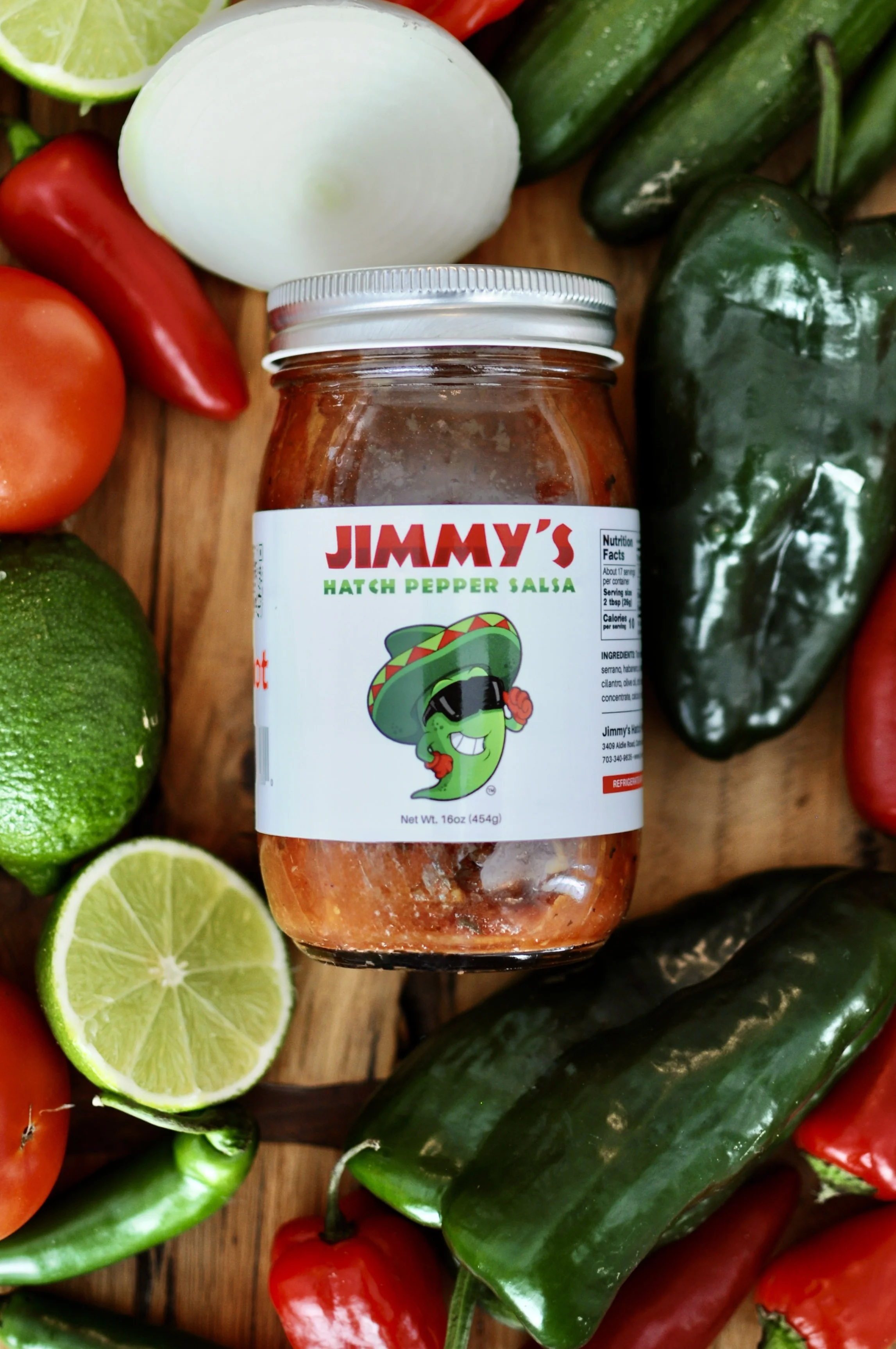 A jar of Jimmy's Hatch Pepper Salsa surrounded by various fresh vegetables including a lime half, onion half, tomatoes, and jalapeño and poblano peppers on a wooden surface.