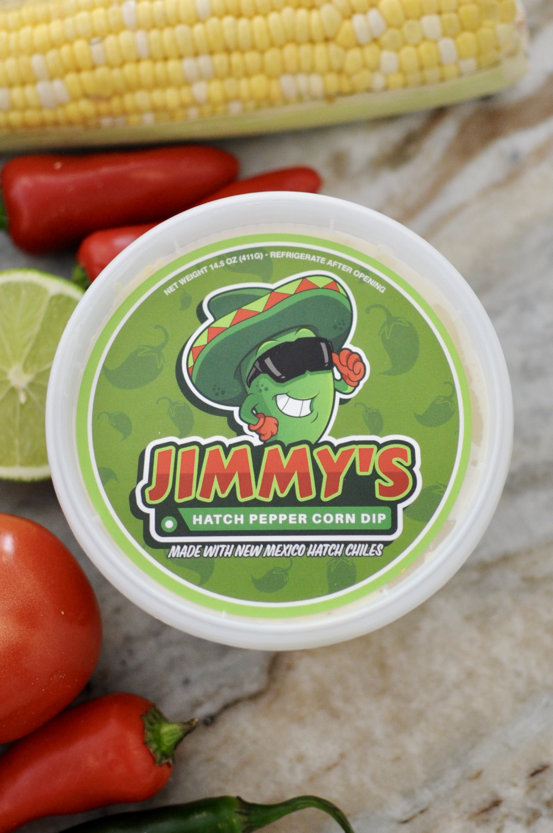 Jimmy's Hatch Pepper Corn Dip, surrounded by fresh ingredients including tomatoes, lime, a jalapeño, and corn.