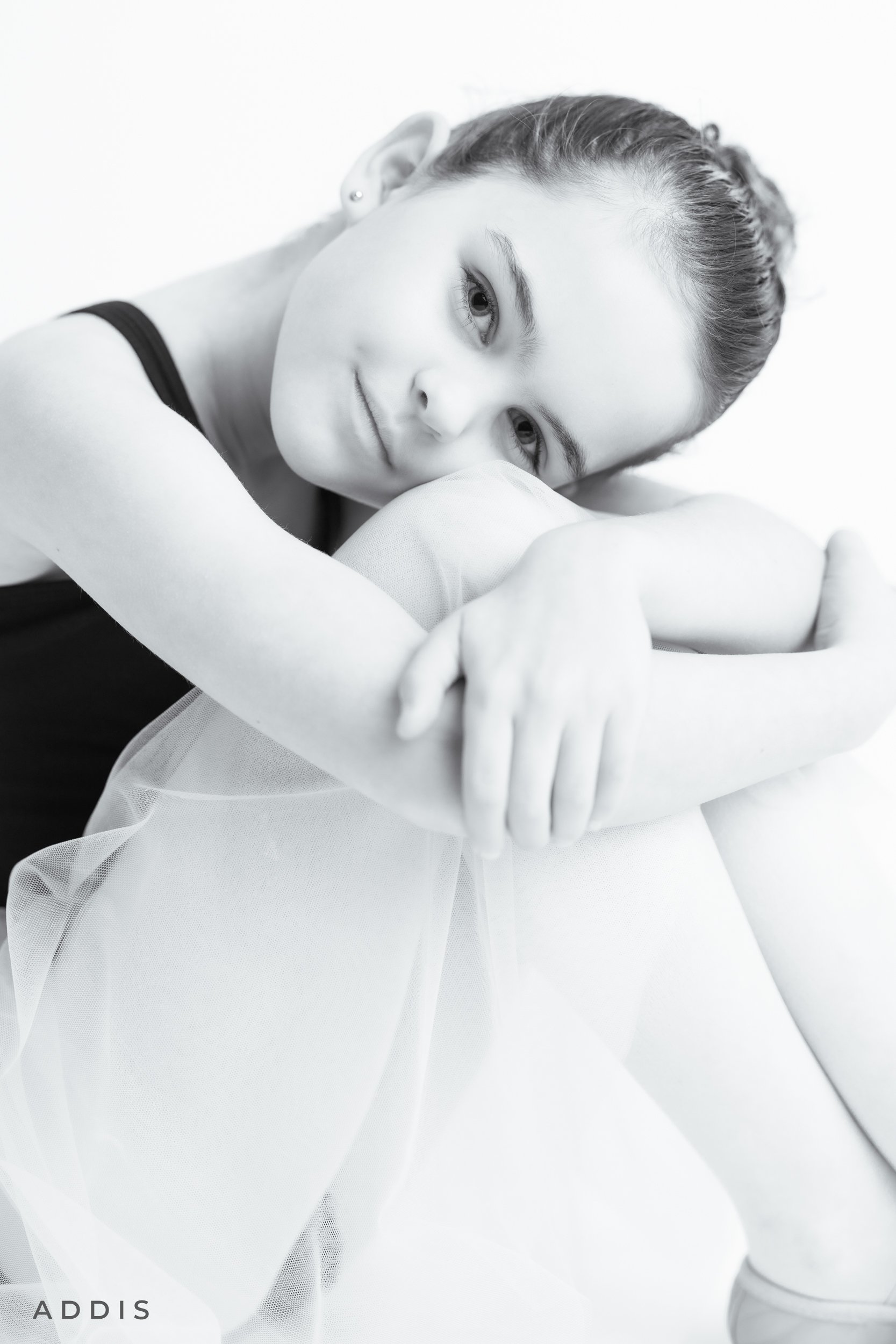 A young woman sitting with her knees pulled up to her chest, resting her head on her knees, looking softly at the camera in a black-and-white portrait.