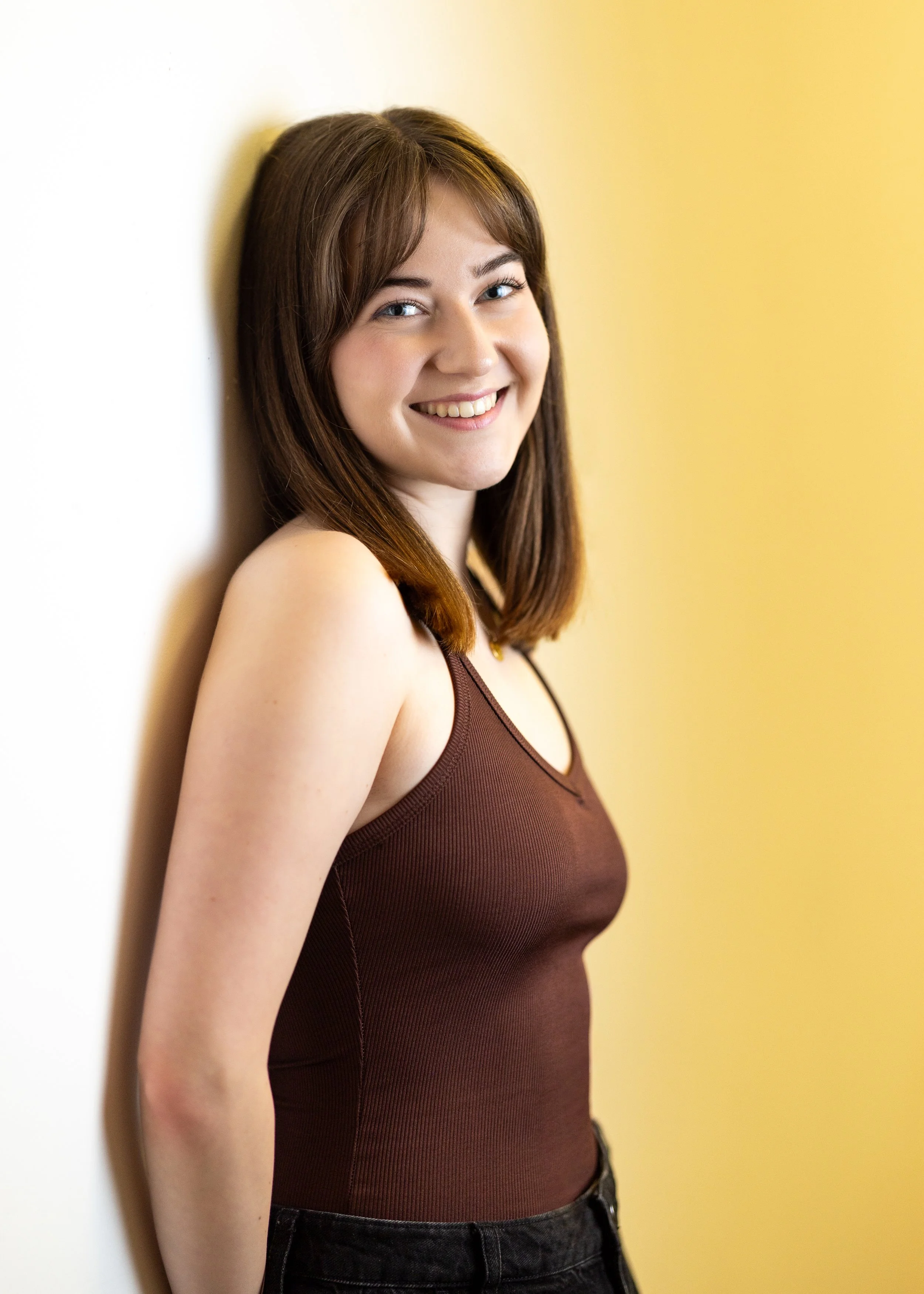 A young woman with shoulder-length brown hair, wearing a brown sleeveless top and black pants, smiling and leaning against a white wall with a yellow background.