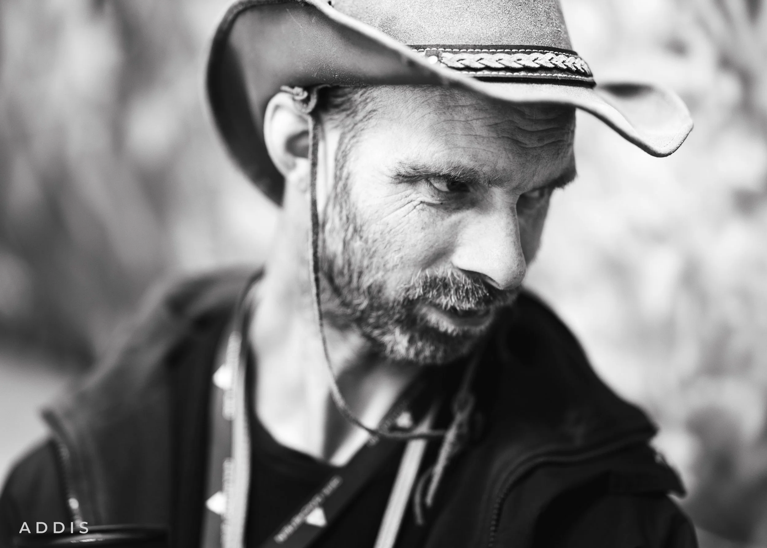 Close-up black and white photo of a man wearing a wide-brimmed hat, looking downward and slightly to the side, with a rugged face and beard.