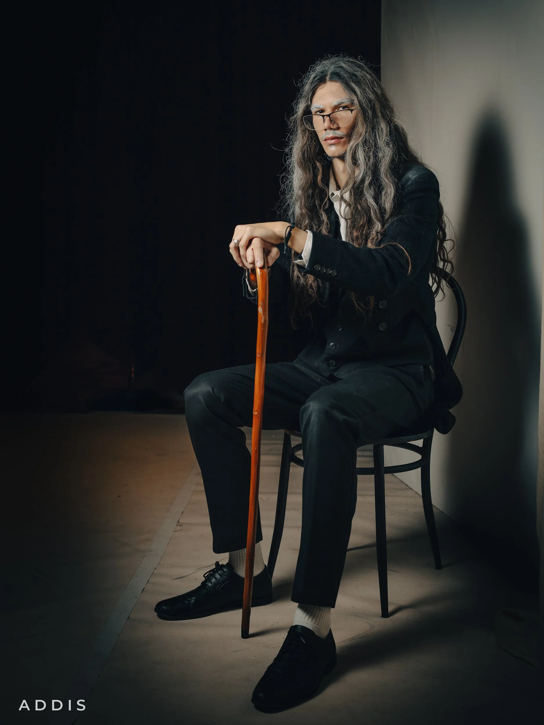 A man from Taming of the Shrew with long gray hair, glasses, and light skin, dressed in a black suit, sitting on a black chair in a dimly lit room with a dark background, holding a wooden cane, looking directly at the camera.