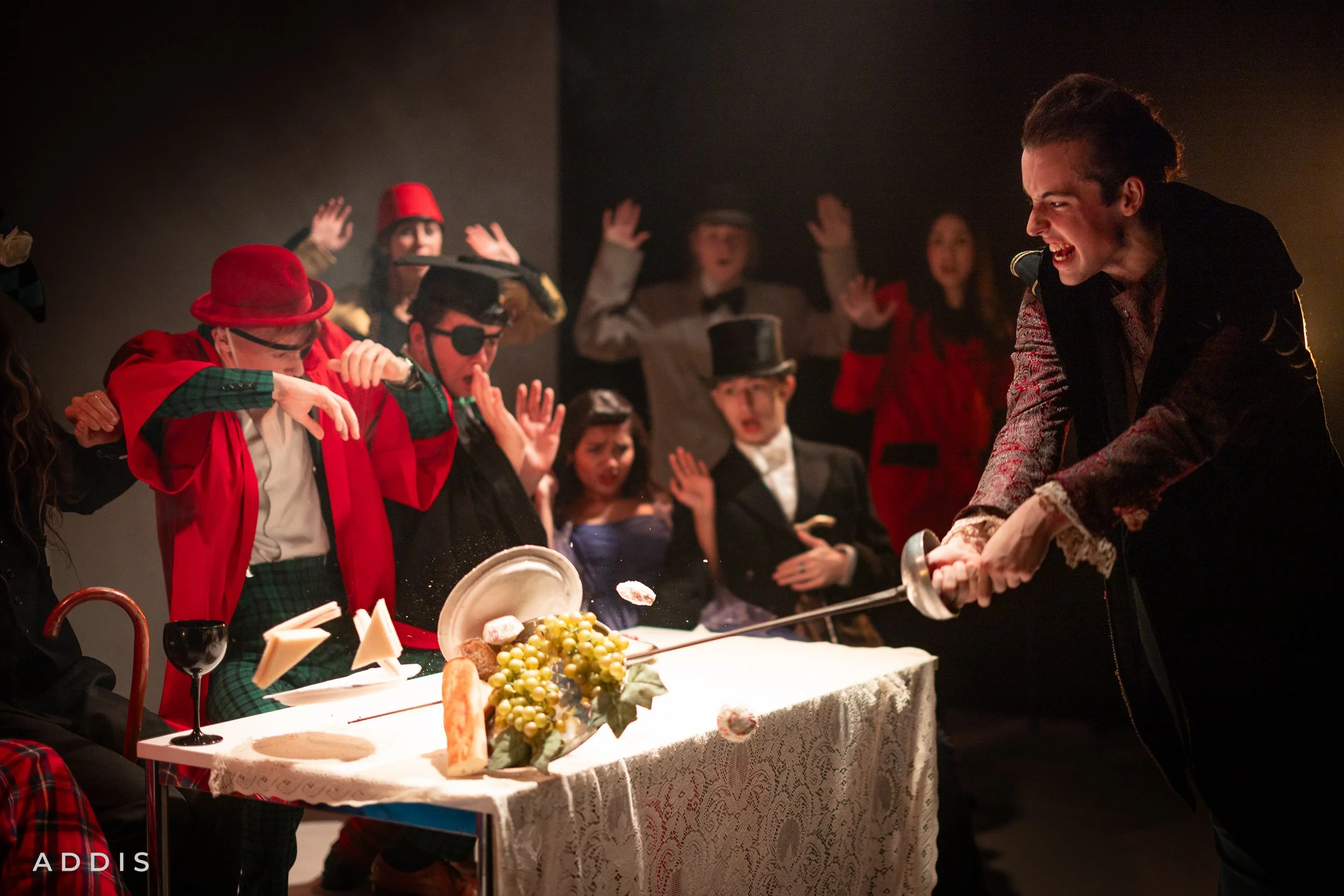 People dressed in costumes, including a person with an eye patch and a person with a red hat, celebrating around a table set with cheese, grapes, and bread while a woman bakes with a pan and others cheer.