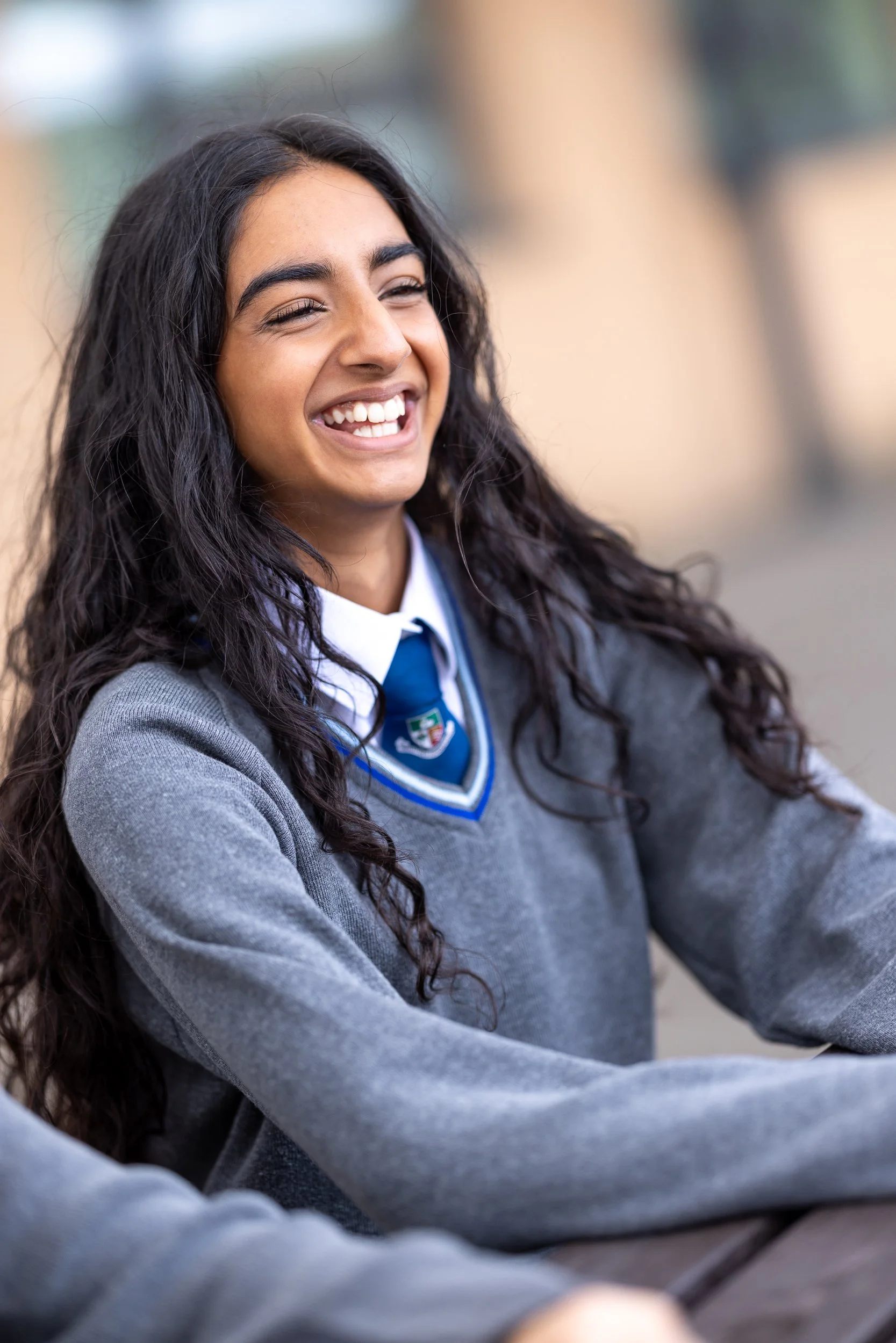 Smiling young woman with long curly hair, wearing a school uniform with a grey sweater and a blue tie, sitting outdoors at The Matthew Arnold School.