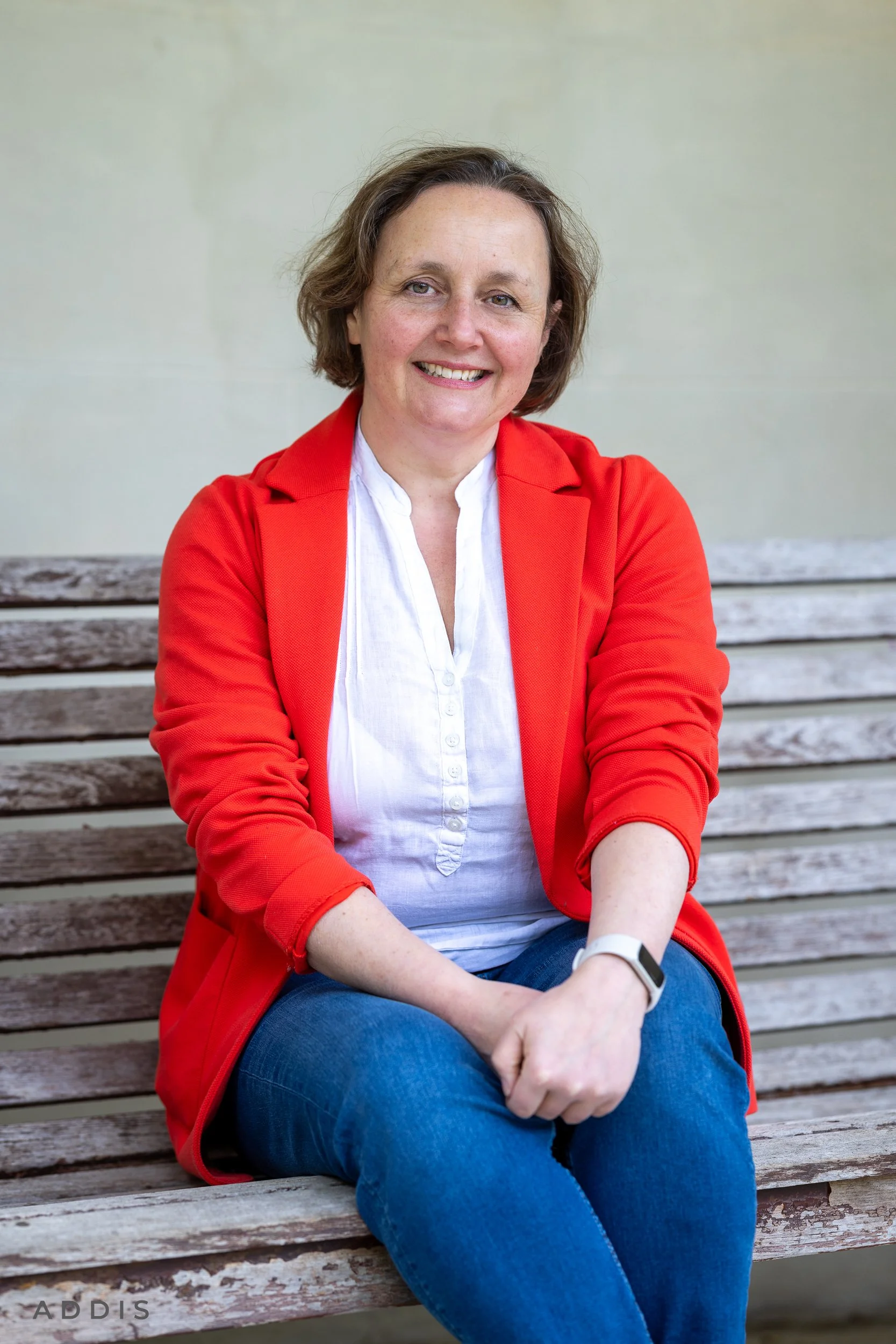 A woman with short brown hair, wearing a red jacket over a white shirt and blue jeans, sitting on a wooden park bench with her arms crossed and smiling at the camera.
