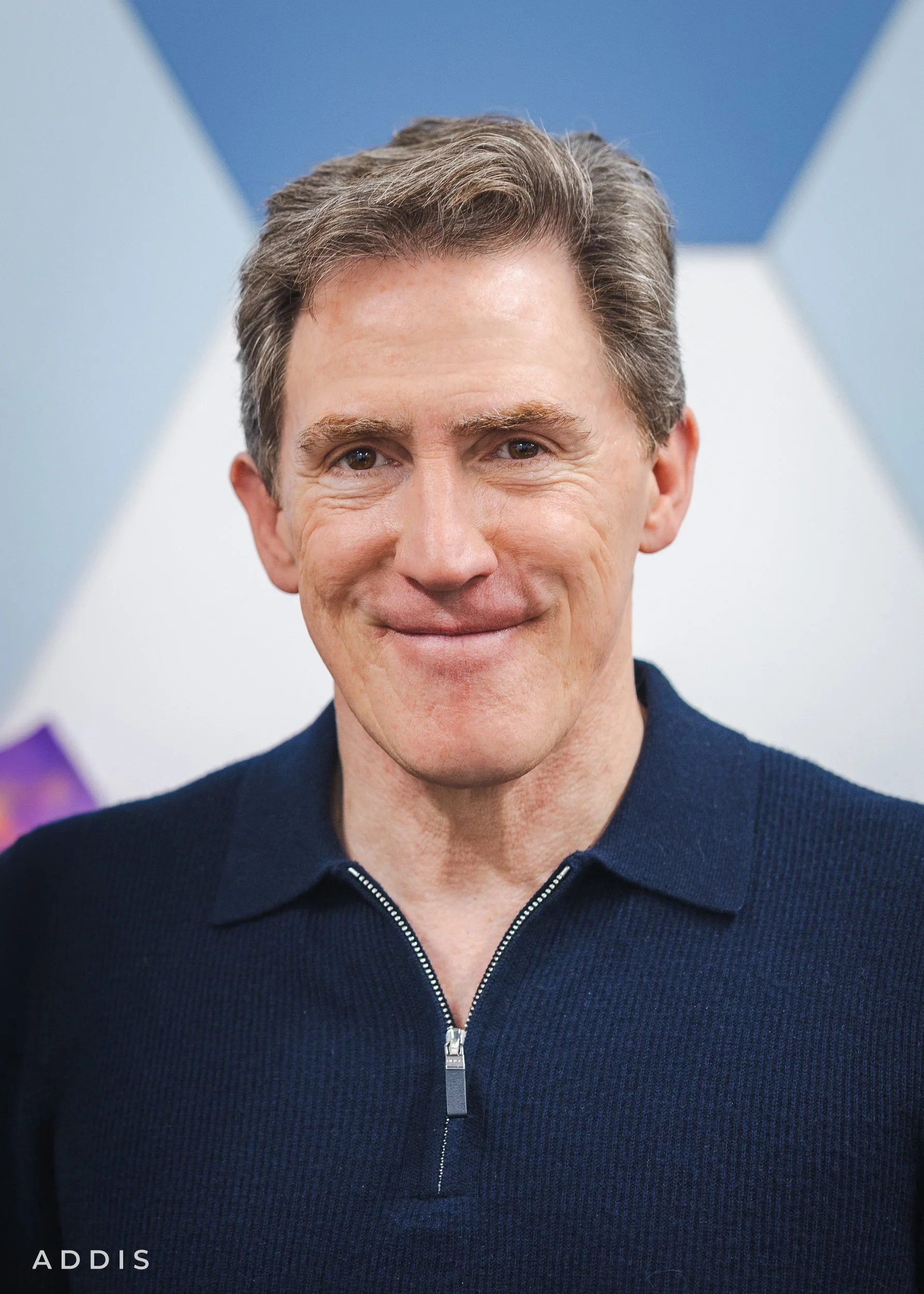 Close-up of a smiling man with short brown hair, wearing a navy zip-up sweater, standing in front of a blue and white background with geometric design.