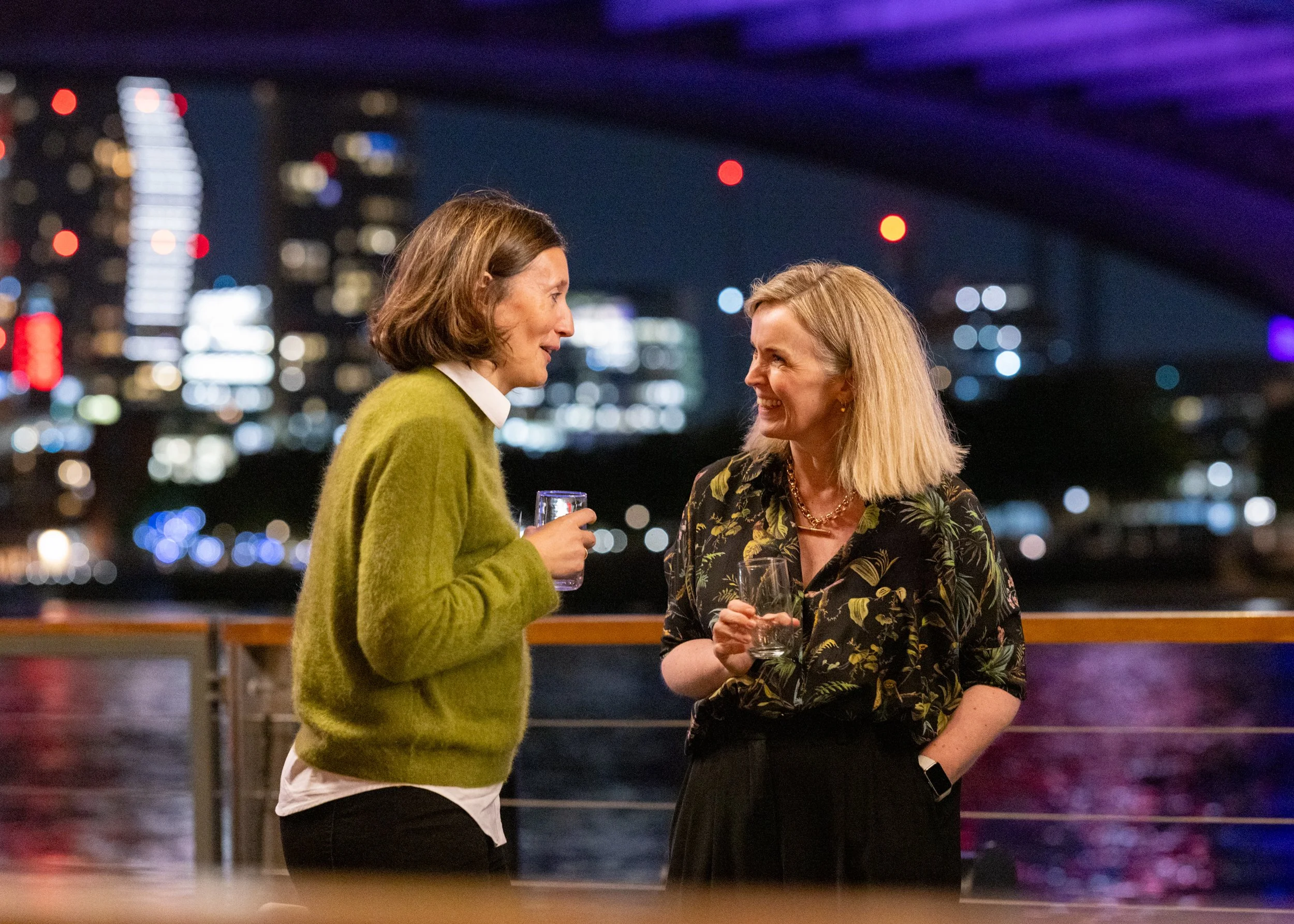 Two women engaging in conversation while holding glasses of drinks at an outdoor gathering during nighttime, with city lights and a bridge in the background.