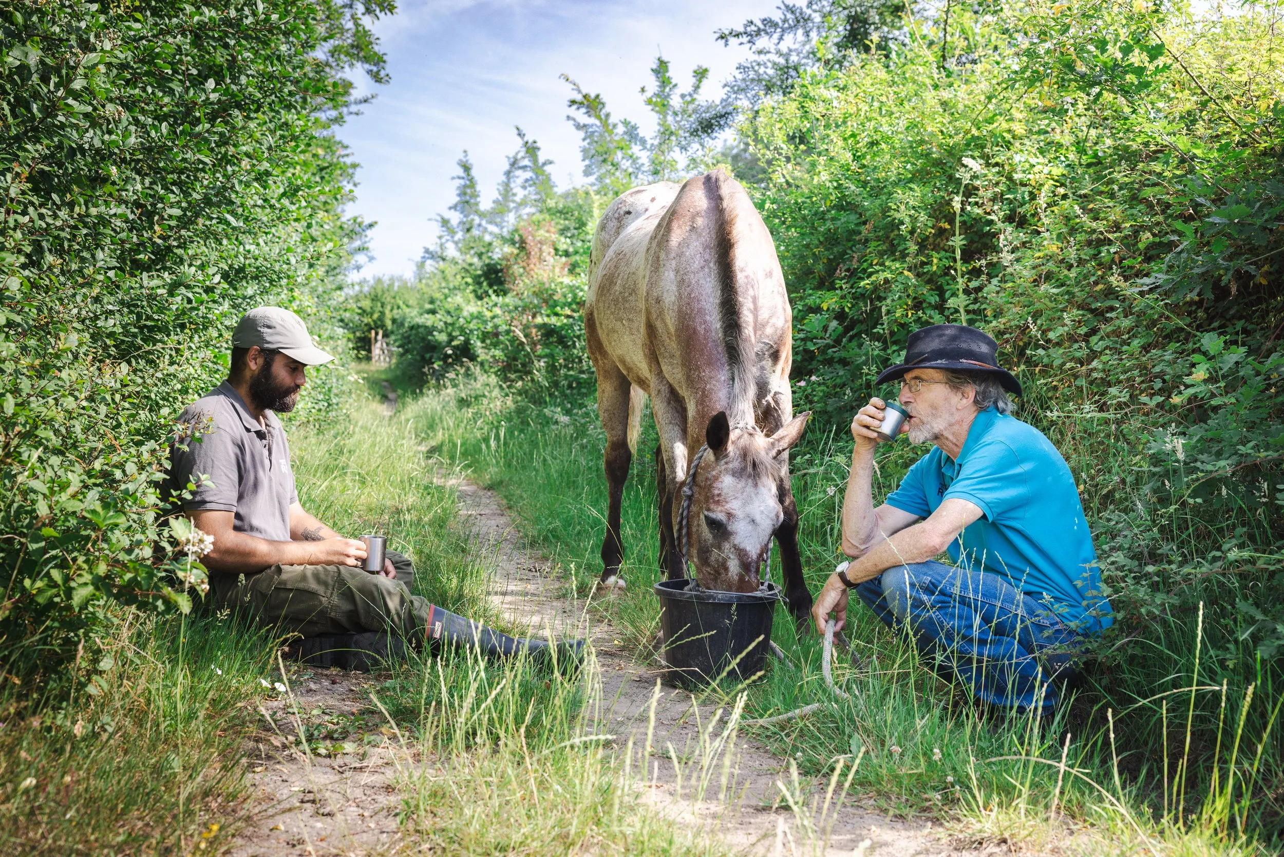 Two men sitting on grass in a green, leafy outdoor area, with a horse drinking from a bucket between them. One man is drinking from a cup, and the other is looking down at his cup. The scene is bright with a clear sky in the background.