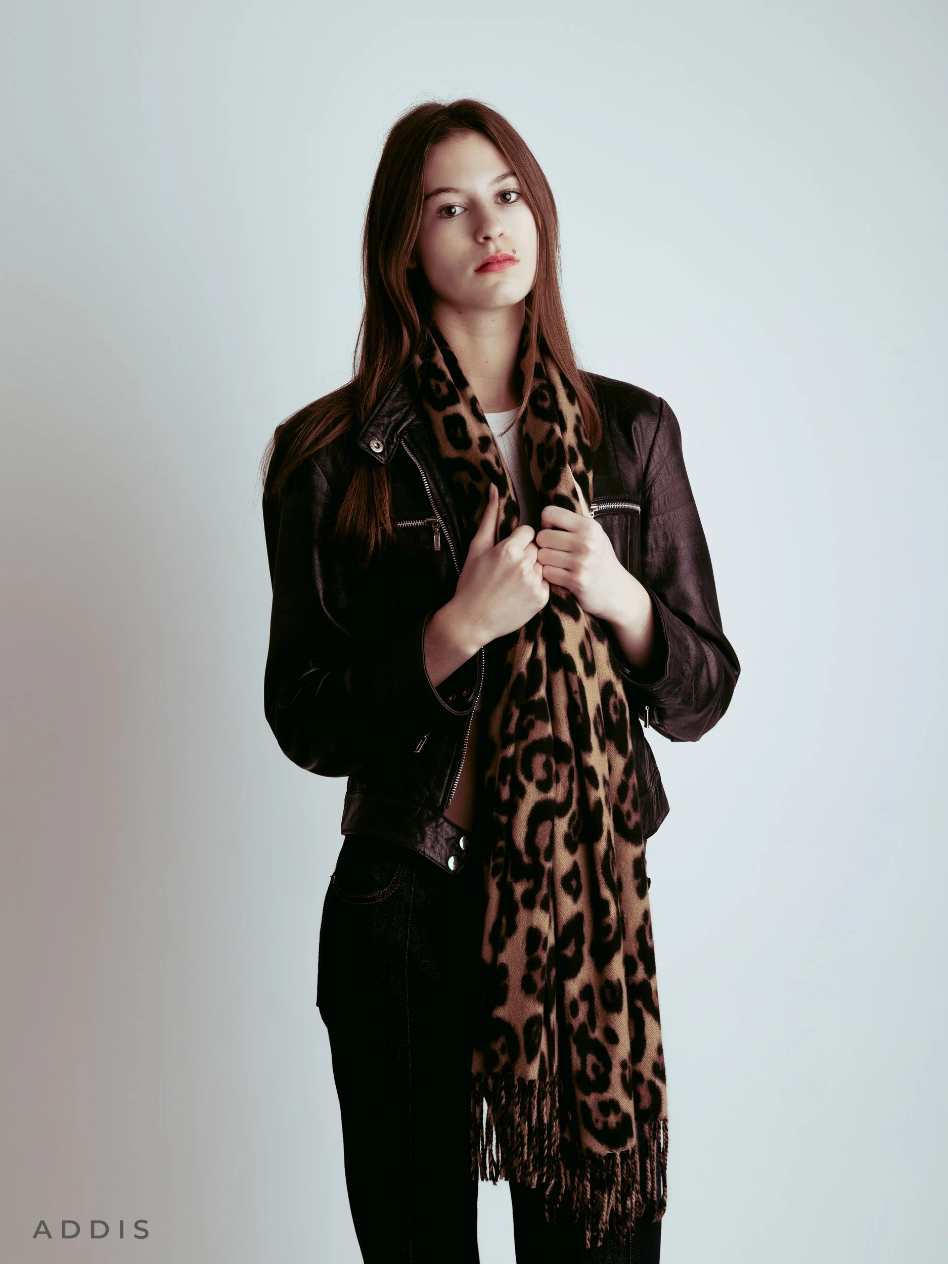 A young woman with long reddish-brown hair wearing a black leather jacket and a leopard print scarf standing against a plain light-colored background.