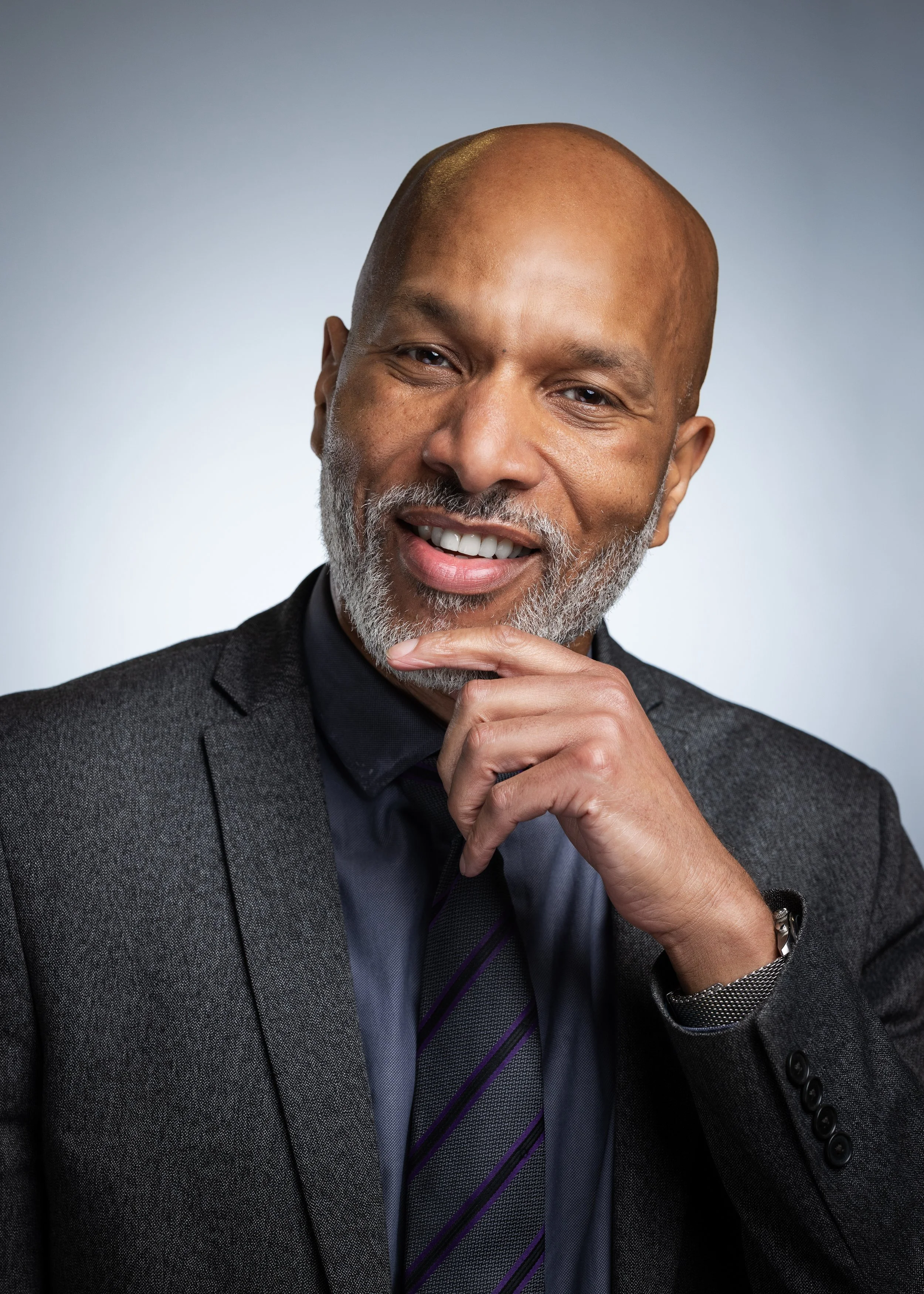 A smiling African American man with a gray beard, dressed in a dark suit with a black shirt and striped purple tie, posing with his hand on his chin against a gray background.
