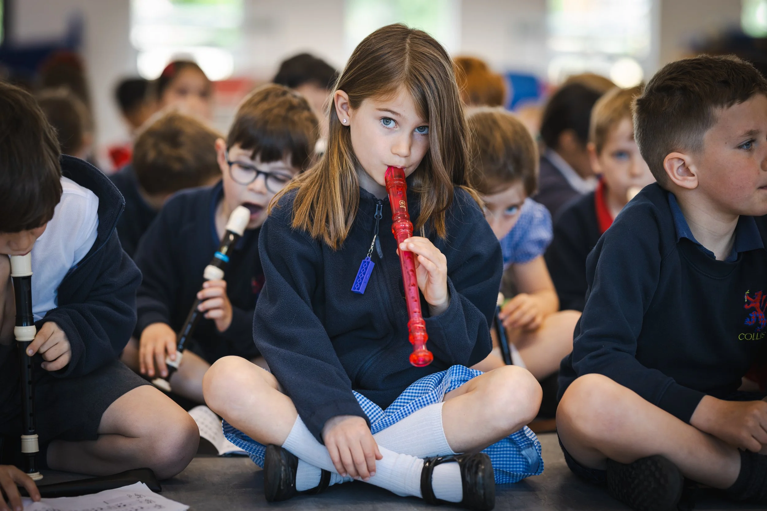 Young girl with blue eyes sitting cross-legged on the floor, playing a red recorder, surrounded by other children, some playing recorders, in a classroom setting.