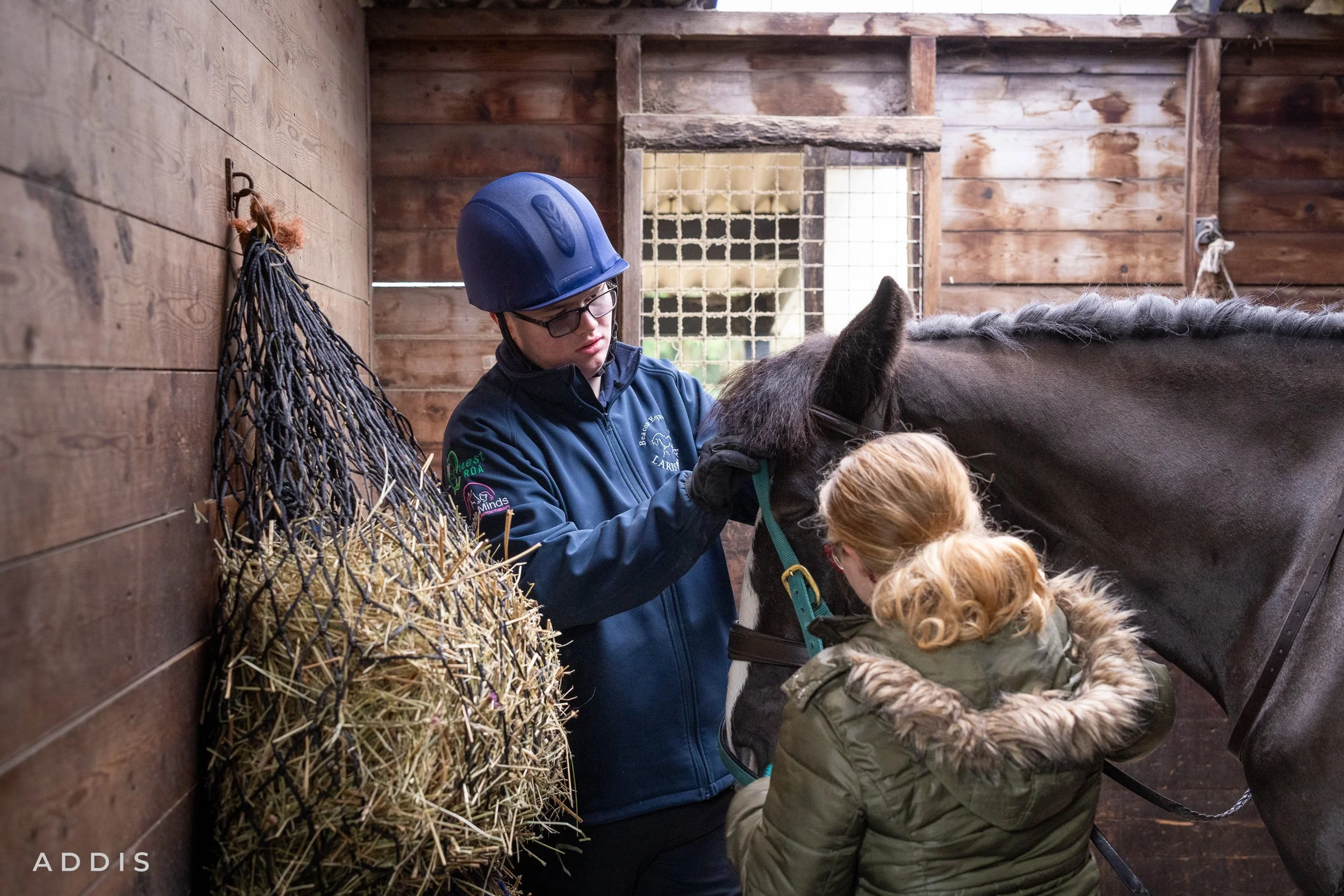 Two children, one wearing a blue helmet, help a woman to put a halter on a horse inside a wooden stable. There is hay hanging on the wall on the left side and a window in the background.