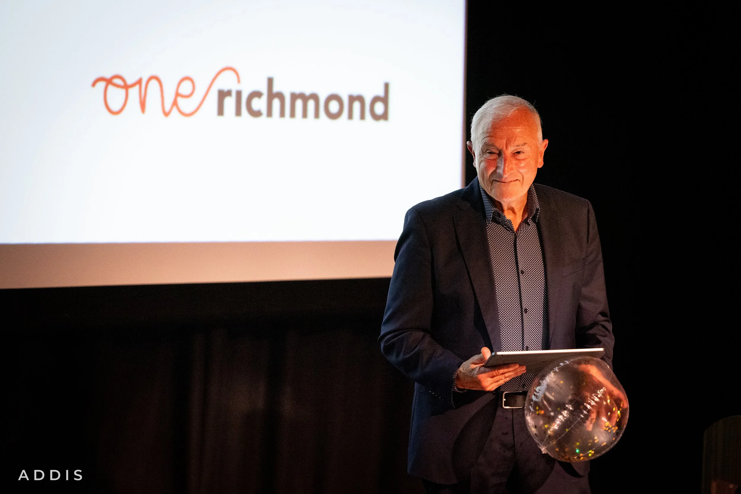 An older man in a suit holding a transparent balloon with colorful confetti inside, standing in front of a large screen displaying a logo with the words 'one Richmond' and a person speaking.