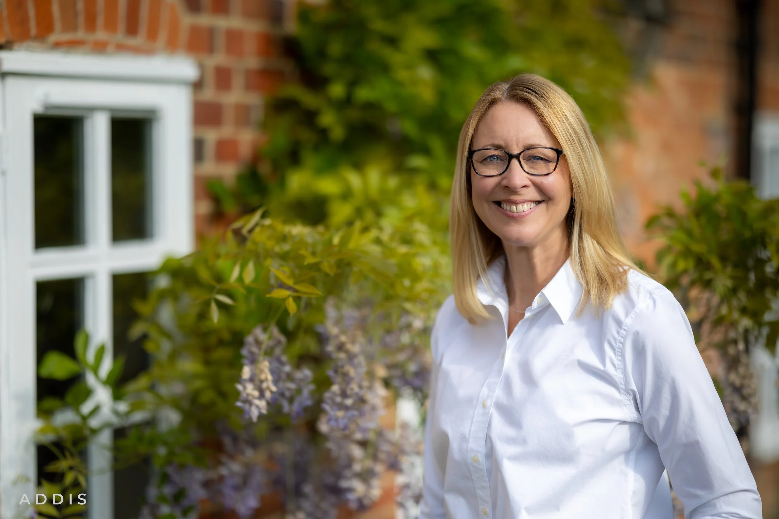 Smiling woman with blonde hair and glasses wearing a white shirt standing outdoors near a brick wall and greenery.