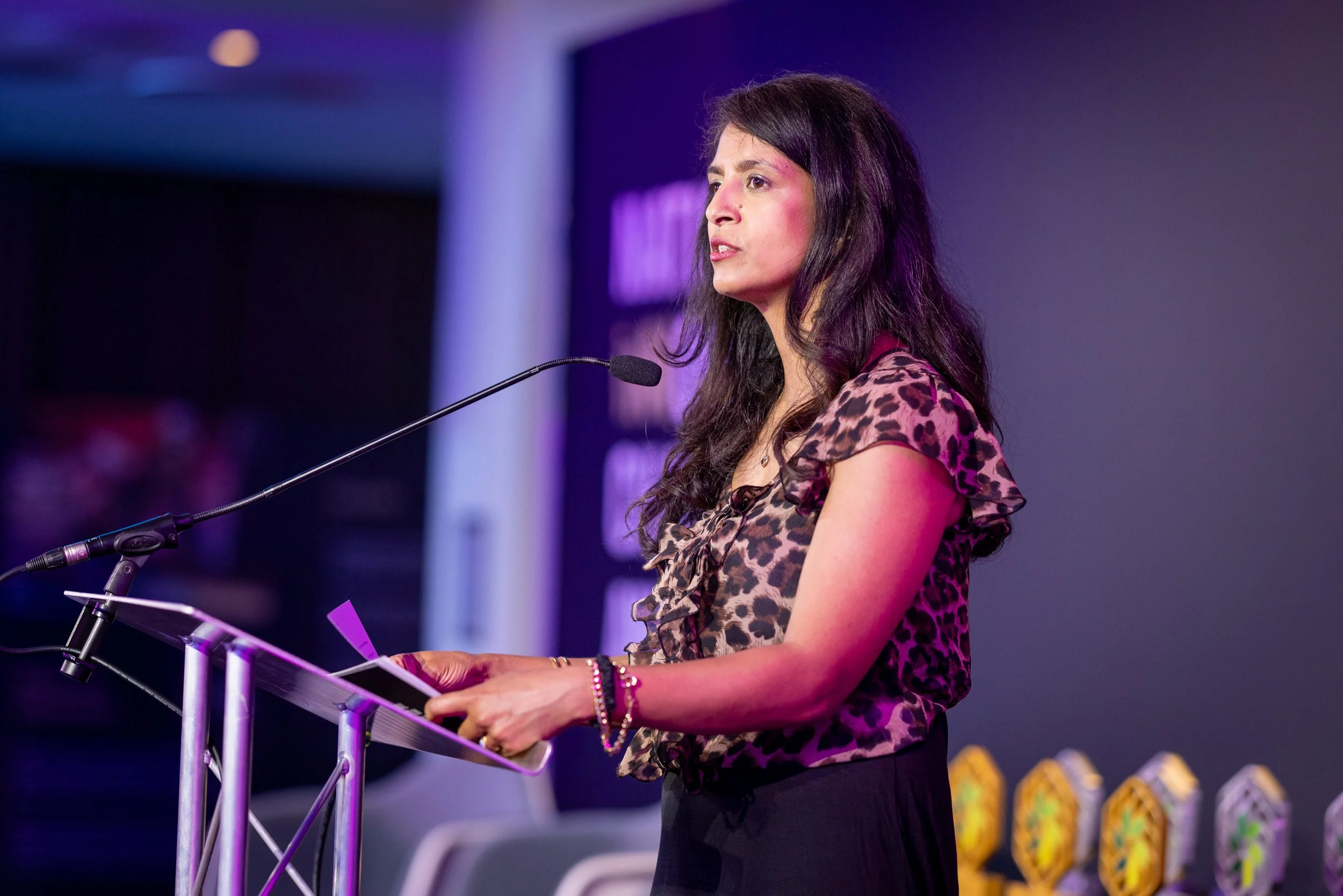 Konnie Huq wearing a leopard print blouse speaking at a podium with a microphone in front of a dark background.
