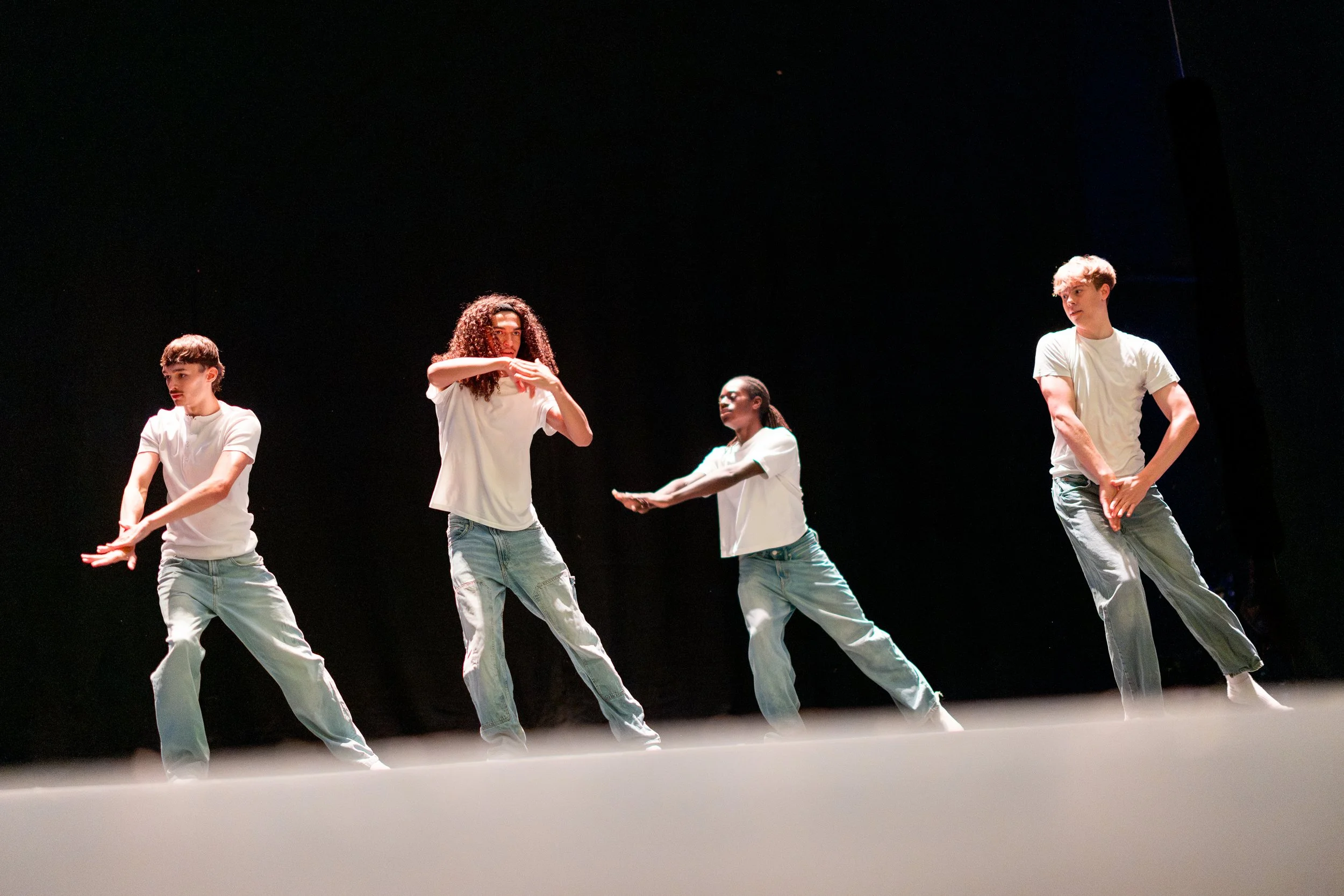 Four young dancers performing on Richmond Theatre stage, wearing casual white T-shirts and jeans, against a black background.