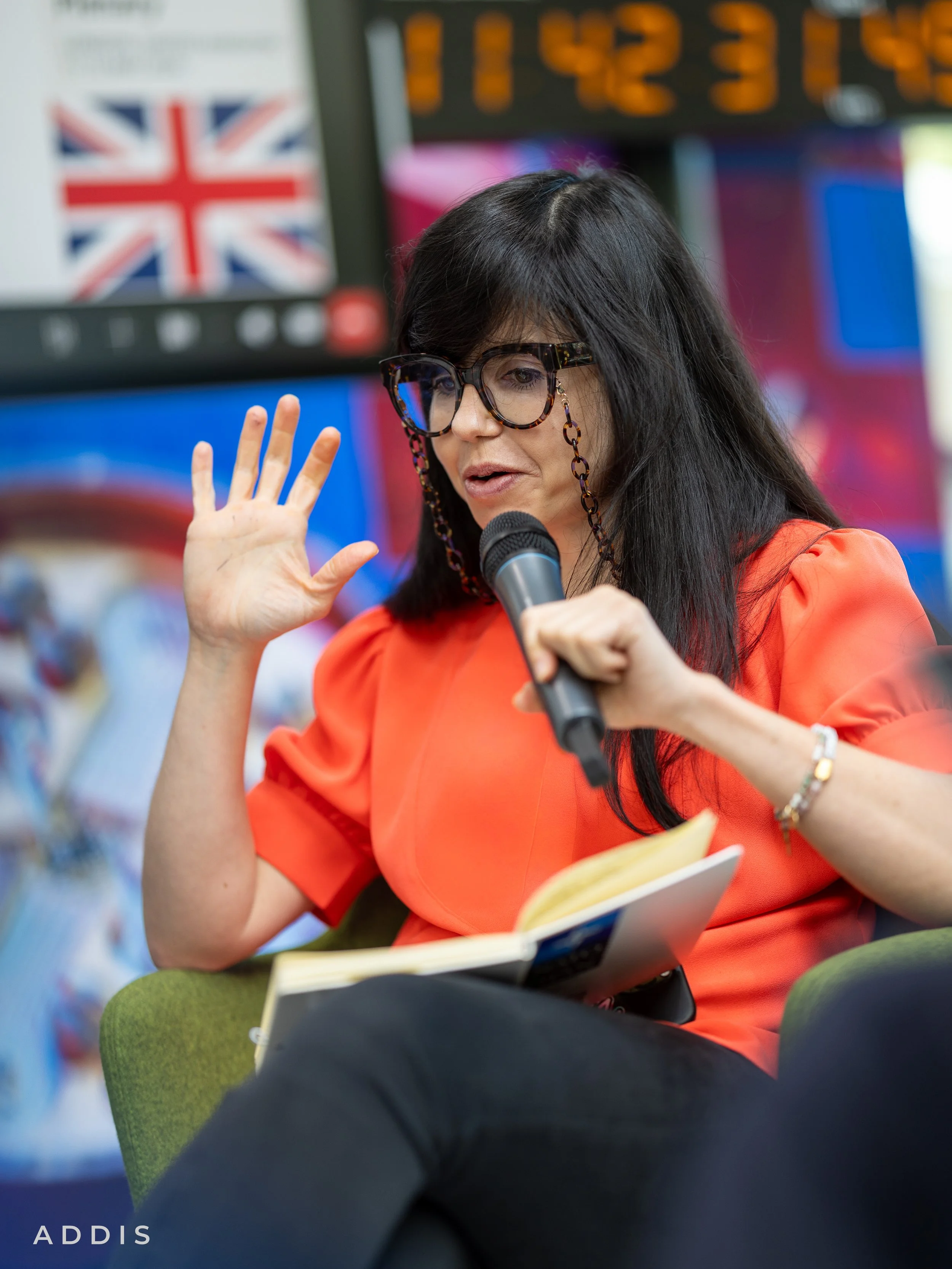 A woman with long black hair, wearing glasses and an orange top, is speaking into a microphone while reading a book. She is sitting on a green chair in a room with colorful background, including a digital display and a blurred British flag.